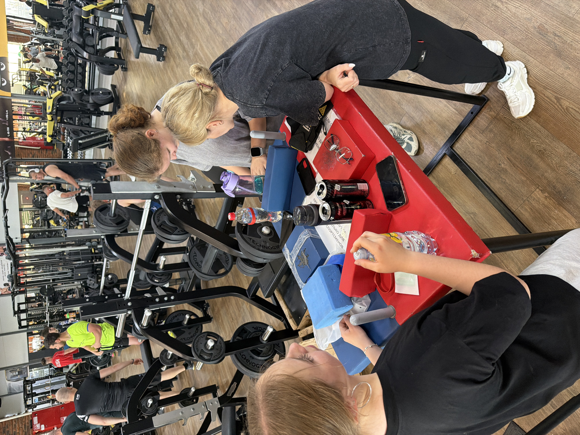 This photograph depicts a group of people in what appears to be a gym setting, situated in the city of Chaikovsky, Russia. The gym is equipped with various weightlifting and exercise machines. The individuals are engaging in activities such as weightlifting and seemingly taking a break or interacting near a table laden with personal items, including bottles, cans, phones, and glasses. The environment is bright and well-lit, suggestive of an indoor facility, and the flooring has a wood-like appearance.