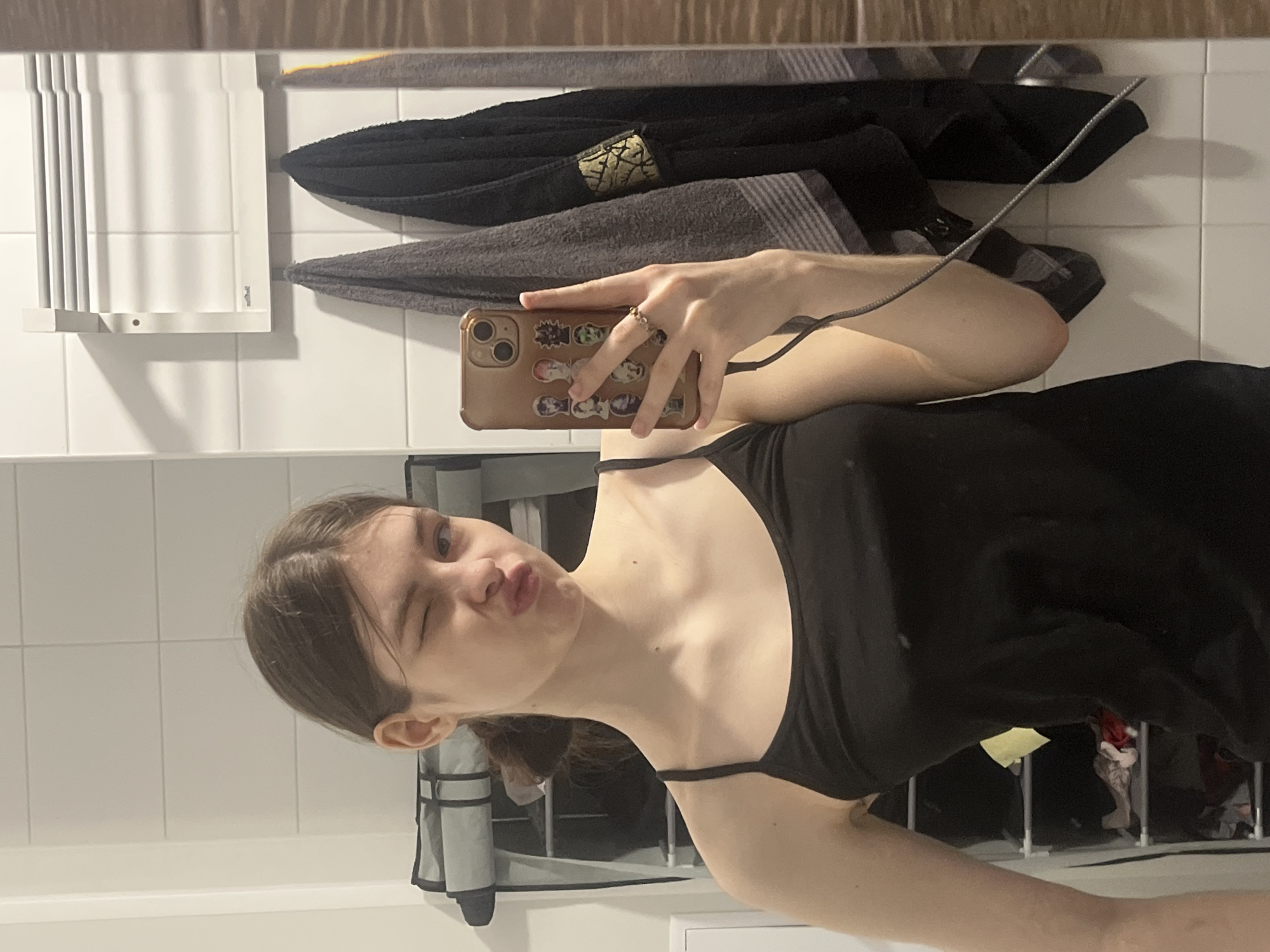 A woman is taking a selfie in a bathroom. The walls are tiled, and there is a towel rack with towels hanging on it. The woman is wearing a black tank top. She is winking and making a duck face. The woman's phone has a brown case with stickers on it. There is a laundry rack in the background. The location in the city of Saint Petersburg, Russia is unknown.