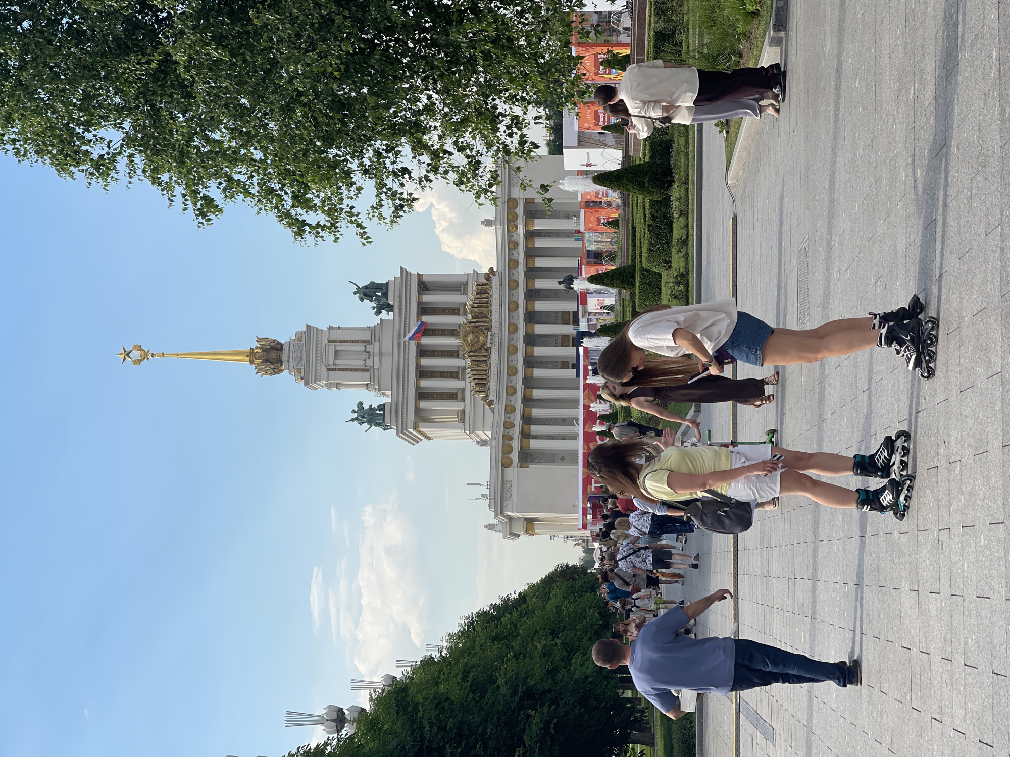 An outdoor scene features a grand building with classical architecture, topped with a golden spire adorned with a star. In front of the building, a pathway is lined with manicured hedges, fountains, and display booths. A large number of people are walking along the pathway, some are wearing casual clothing, while two women in the foreground are wearing rollerblades. The sky is blue with scattered clouds, and trees with lush green foliage line the left side of the pathway.
