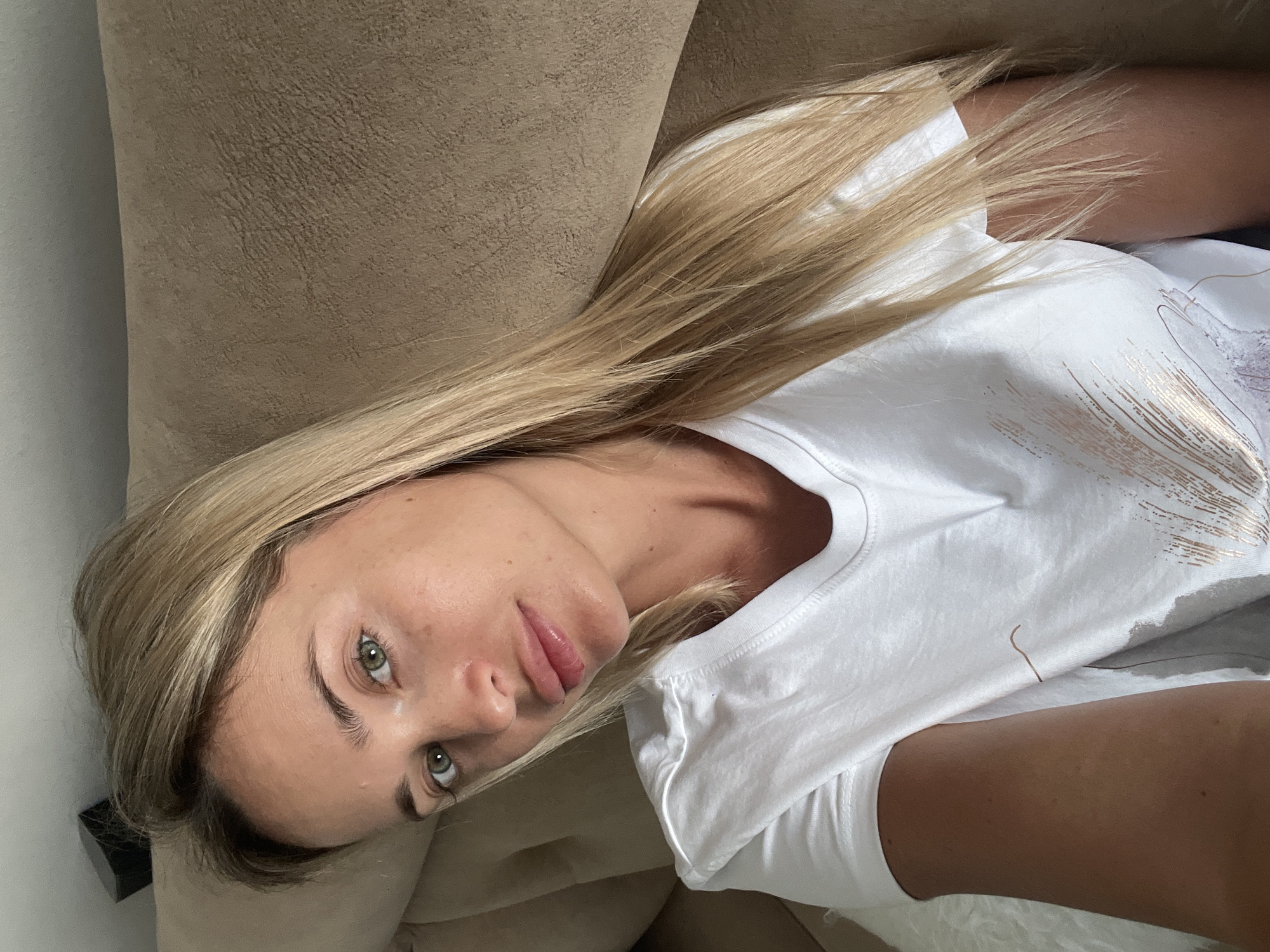A woman with long blonde hair and green eyes is photographed from the chest up, reclining on a beige sofa. She is wearing a white t-shirt with a metallic print. The background appears to be an indoor setting, possibly a hotel room in Sharm El Sheikh, Egypt. The lighting is natural and soft, suggesting daylight.