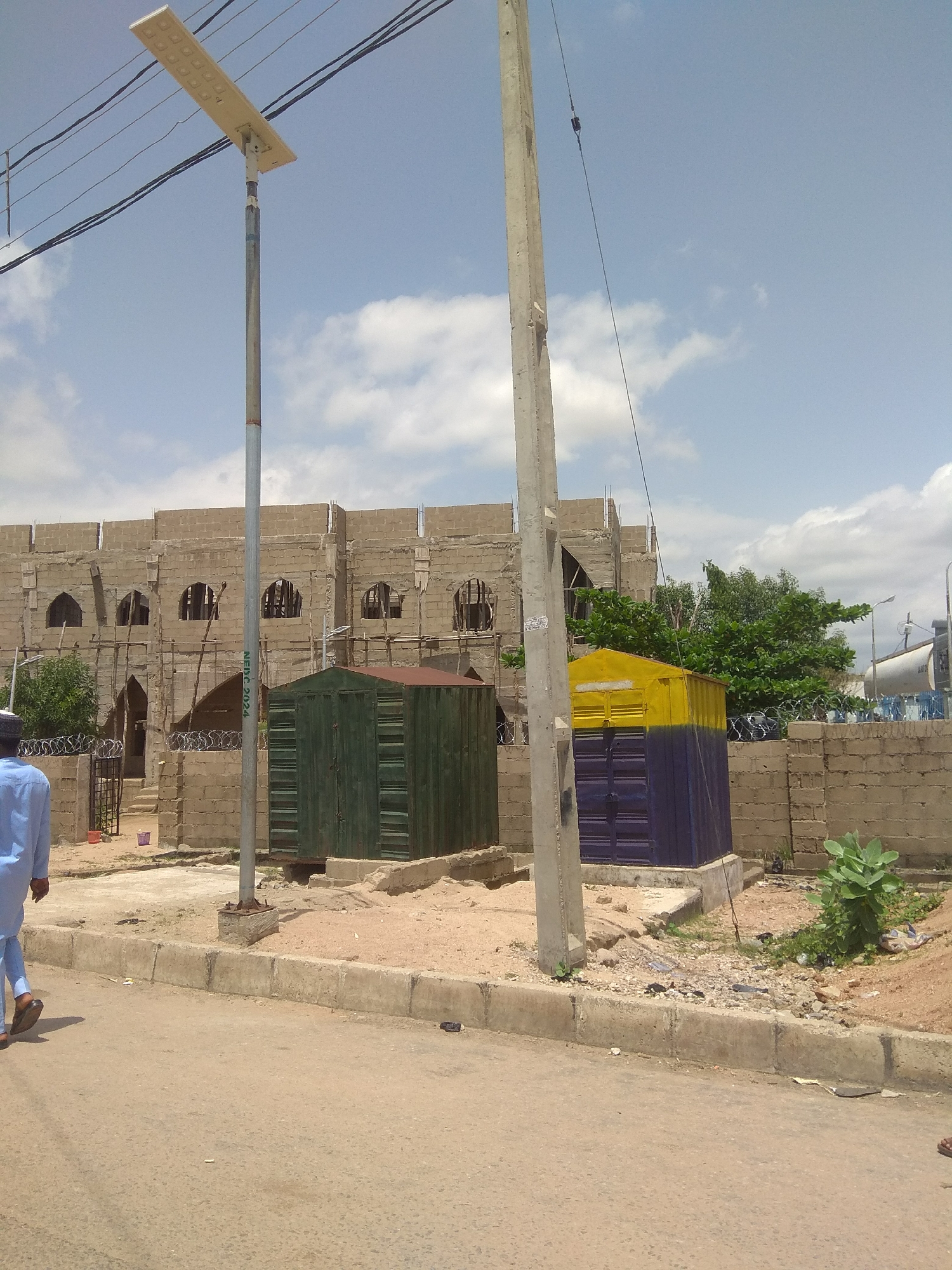 The image depicts an outdoor scene in Jalingo, Nigeria, under a bright, partly cloudy sky. The time of day appears to be midday. The primary subjects are a partially constructed building made of tan bricks, two small utility sheds, and a person walking away from the camera.
The building, which has arched window openings and scaffolding, suggests ongoing construction. One shed is painted dark green, and the other is a two-toned structure with a yellow top and a dark blue bottom. Both sheds are rectangular and appear to be made of metal. Razor wire is visible along the top of a brick wall surrounding the property.
A person dressed in a light blue traditional outfit is walking on the left side of the image, on a dirt and paved area near a street. They are walking away from the viewer, making it impossible to discern their facial features or specific activity beyond walking.
Other objects visible include a solar-powered street lamp on a tall pole, a concrete utility pole with some wires attached, and some sparse vegetation. Debris and uneven ground are present around the utility structures and the construction site, indicating a somewhat unkempt environment. The overall scene suggests a developing or under-construction area in a town or city. No specific event is clearly depicted, other than the general activity of construction and the presence of people.