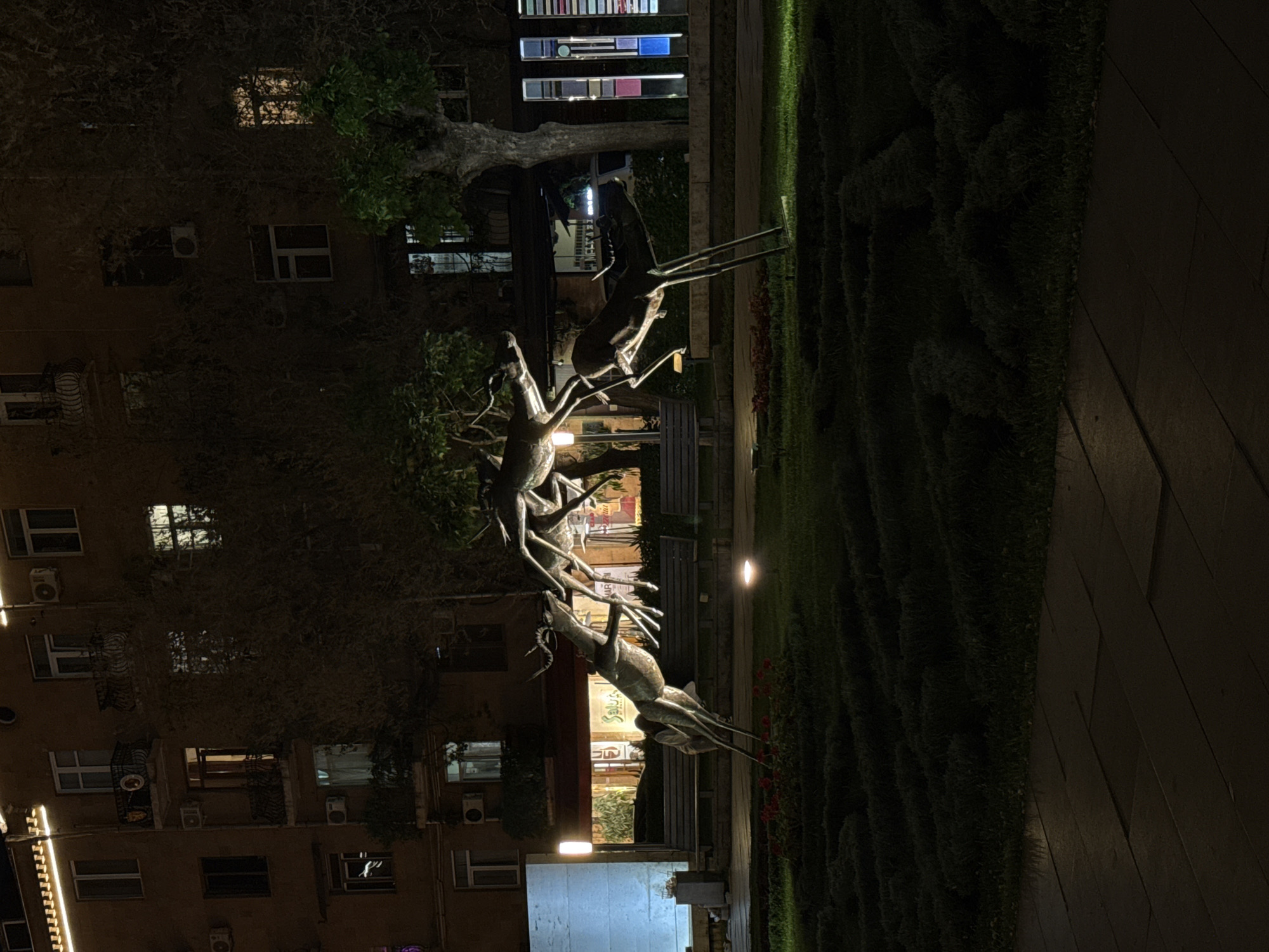 The image shows a nighttime view of a sculpture in a park or plaza, possibly in Unknown, Unknown, given the unknown location. The sculpture depicts a group of stylized animals, possibly deer or similar creatures, appearing to climb or interact with each other. The sculpture is metallic and stands on a grassy area bordered by a stone or paved walkway. In the background, there is a multi-story building with windows and air conditioning units visible on its facade. The scene is illuminated by artificial lights, including streetlights and lights emanating from the building, creating a contrast between light and shadow. There are benches and greenery in the vicinity of the sculpture, suggesting a public park or gathering space.