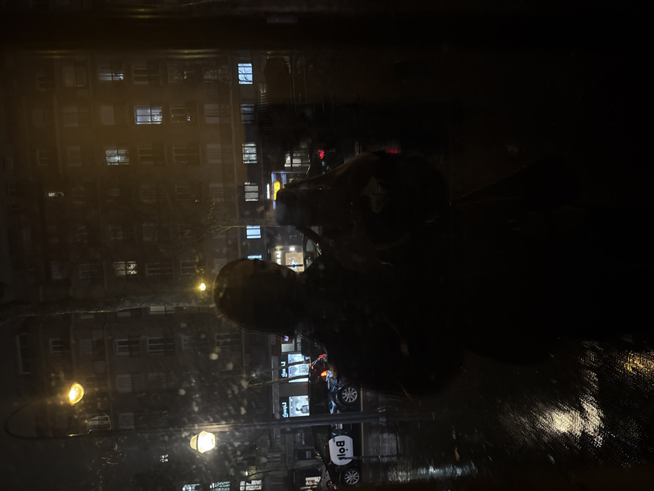 Nighttime scene, likely taken through a rain-streaked window. Buildings with illuminated windows create a hazy, urban backdrop. Streetlights cast a warm glow on the wet pavement. A dark figure, possibly a person in a coat or with a backpack, is silhouetted in the foreground. A "Bolt" car is parked on the street. The overall atmosphere is dark, rainy, and urban.