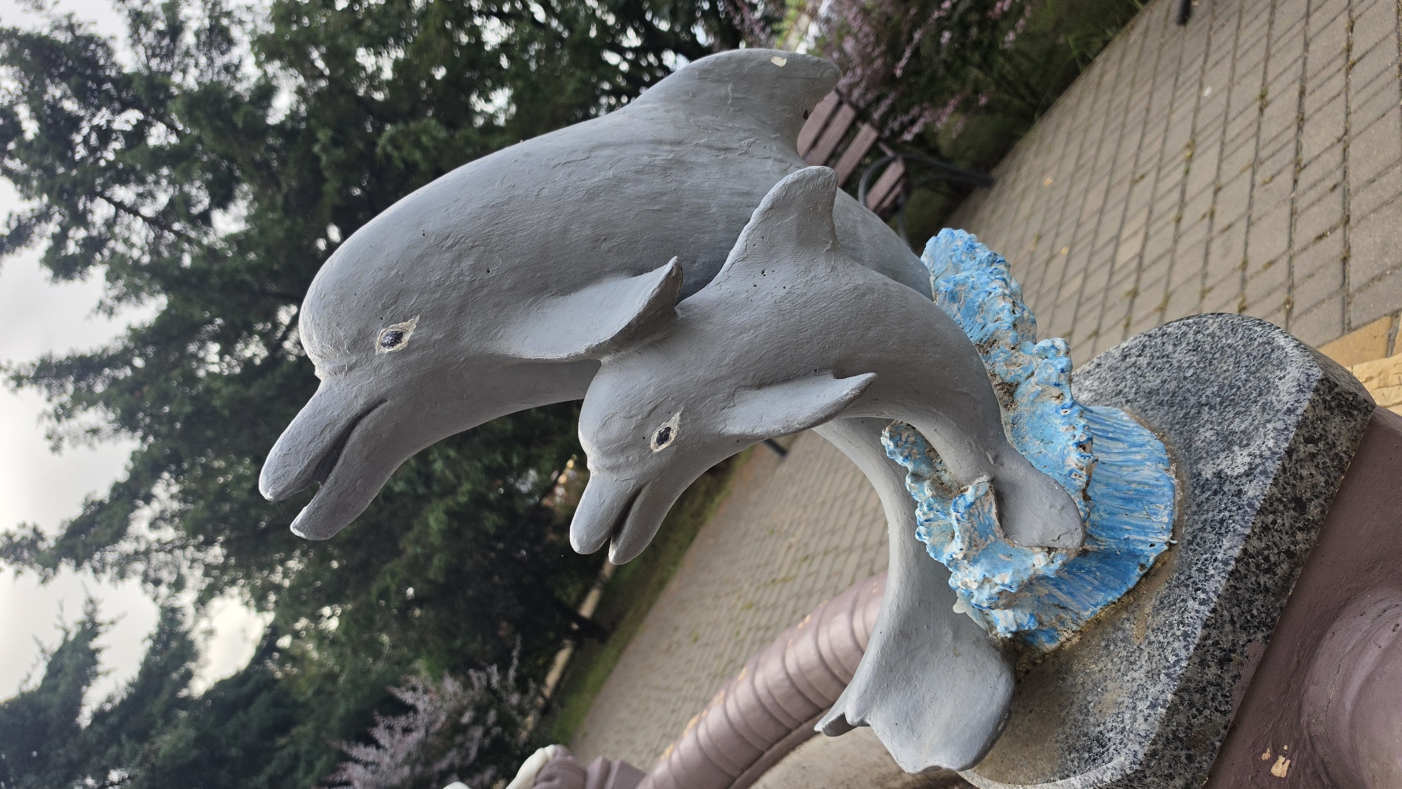 A close-up, low-angle shot shows a gray sculpture of two dolphins appearing to leap out of blue, wave-like forms. The larger dolphin is positioned behind and above the smaller one, both with their mouths open and eyes visible. The sculpture rests on a granite base, which is on top of a larger, purplish-brown base. The background is blurred, but appears to be an outdoor setting with trees, foliage, a paved walkway, and a park bench. The lighting suggests it is daytime, possibly overcast.