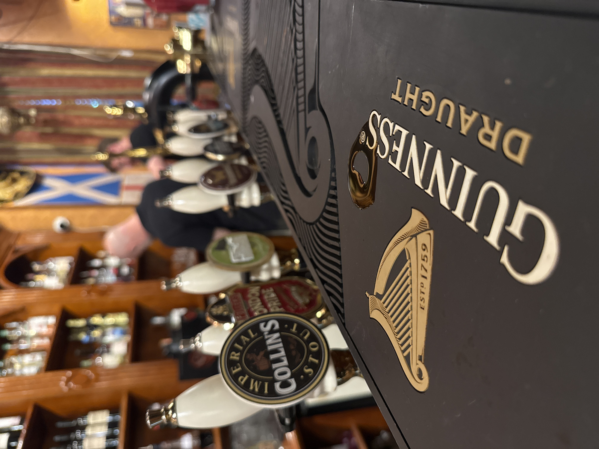 The image shows a close-up view of a bar counter with several beer taps. In the foreground, a Guinness Draft tap handle is prominently displayed, featuring the Guinness harp logo and the text "ESTD 1759". Behind the Guinness tap, other beer tap handles are visible, including one labeled "COLLIN'S STOUT" and another with "LONDON PRIDE". The background reveals shelves stocked with various bottles of alcohol, some people standing at the bar, and a Scottish flag hanging on the wall. The scene suggests a pub or bar setting. The question of the location being in Saint Petersburg, Russia is not supported by any visual evidence in the image.