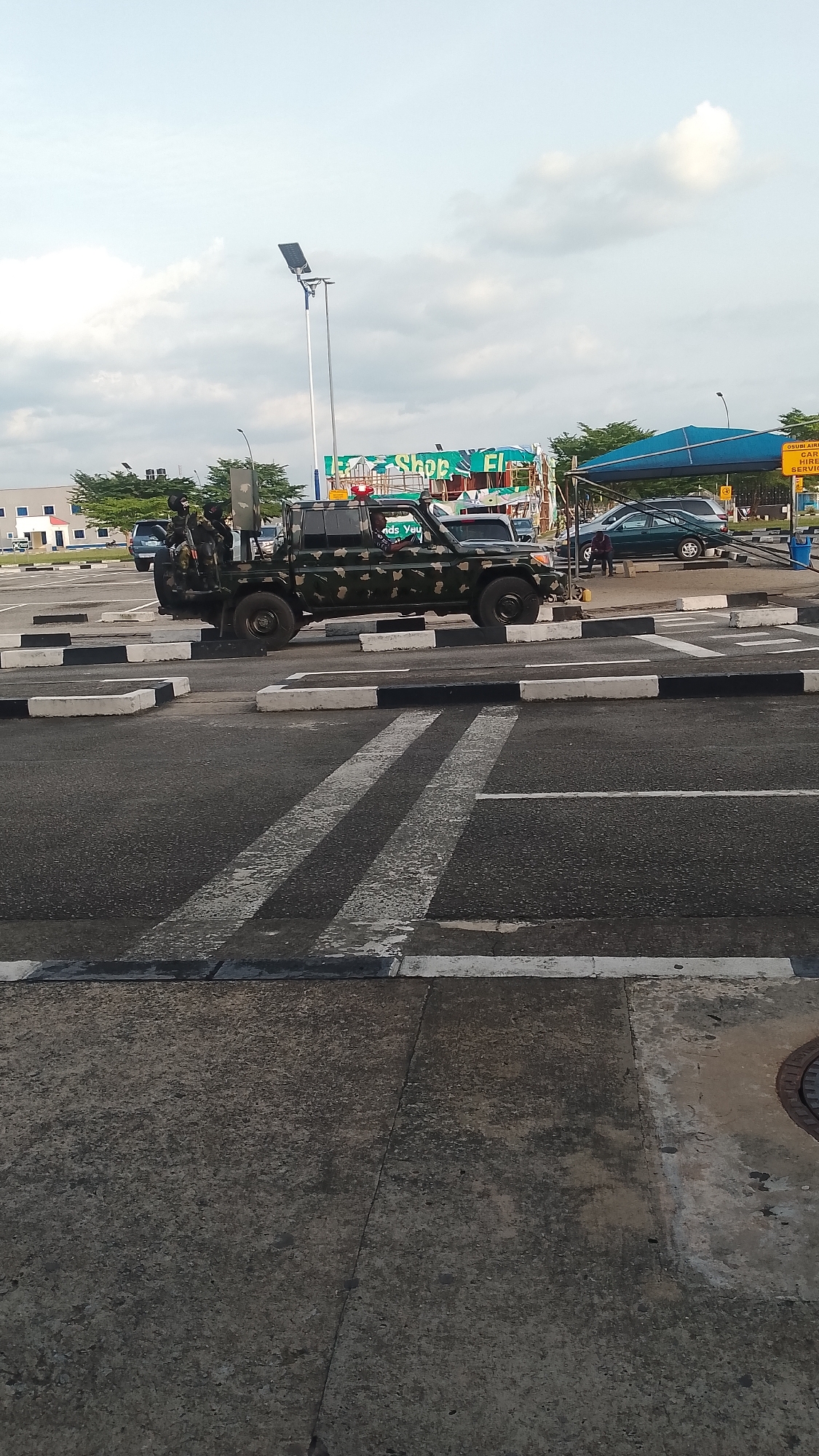 This daytime image captures an outdoor scene in what appears to be a public area in Orerokpe, Nigeria, under an overcast sky. The central subject is a camouflage-painted military-style pickup truck positioned on an asphalt road featuring black and white speed bumps and white zebra crossing lines. Two uniformed individuals, likely military or security personnel, are visible in the truck's rear bed; one stands, appearing to hold a firearm, while another person is in the driver's seat. A red warning light is mounted on the truck's roof.
In the background, to the left, modern, light-colored buildings and green trees are visible. To the right, civilian vehicles, including a dark blue SUV and a green minivan, are parked or moving. A person is seated under a blue canopy near these vehicles. Behind the truck, a green-tarped structure, possibly a market stall or shop, has the text "Shop El" partially visible. A tall street light with a solar panel stands prominently in the mid-ground.
On the far right, a yellow sign displays partial text, "OSUBU AIM," and clearly readable "LAGOS HIRE SERVICE," indicating a transportation service. The overall scene suggests a busy roadside, transport hub, or market area, characterized by both commercial activity and a noticeable security presence, with the personnel appearing watchful.