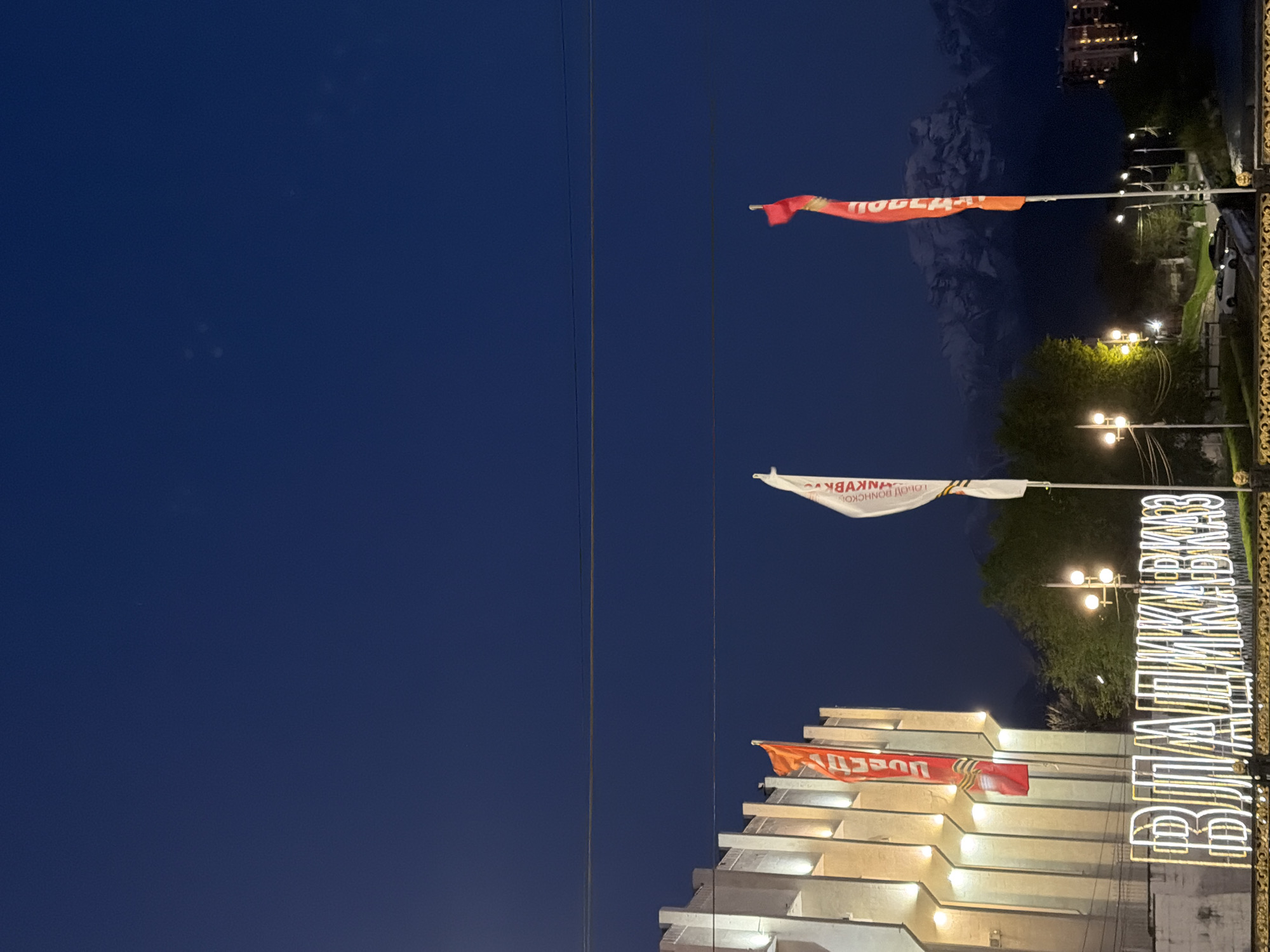 The image shows a nighttime scene with several flags and illuminated signs in the city of Chermen, Russia. In the top-right corner, an orange flag with the word "ПОБЕДА" (Victory) is displayed. Below that, a white flag is seen with the words "СЛАВА ВОИНАМ ДОНСКОЙ ЗЕМЛИ" (Glory to the Warriors of the Don Land). A building with orange flags is located in the lower part of the image, and the sign "ВЕЛИКИМ КИМКОВЦАМ" (To the Great KIM workers) is vertically displayed. Streetlights are visible. The background shows a dark blue sky and outlines of distant mountains. Powerlines run vertically across the image.
