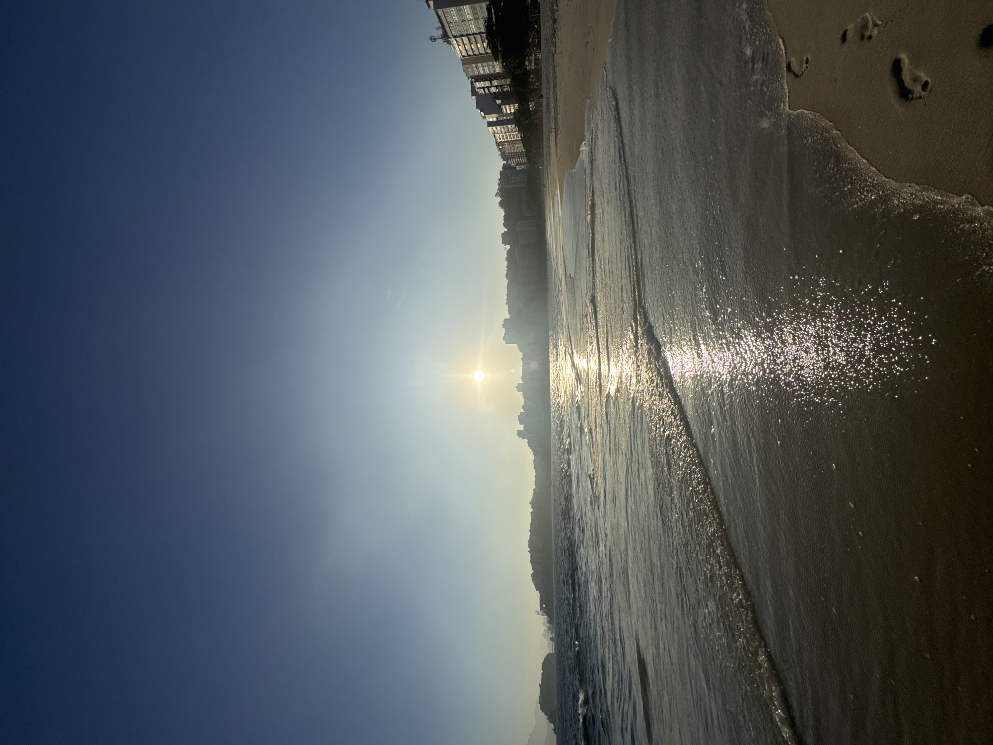 This image captures a serene coastal view at sunrise or sunset, likely from the popular Icaraí beach in Niterói, Brazil. The sun, a bright orb, is positioned centrally in the sky, casting a shimmering path across the calm ocean waters. Gentle waves lap at the sandy shore, leaving a wet sheen and a few footprints. In the background, a line of multi-story buildings, characteristic of a coastal city, stretches along the coastline, with some vegetation visible. A distant, hazy landmass or island is also present on the left side of the horizon. The sky is a clear, gradient blue, transitioning from a deeper hue at the top to a lighter tone near the horizon. The overall scene evokes a peaceful and tranquil atmosphere.