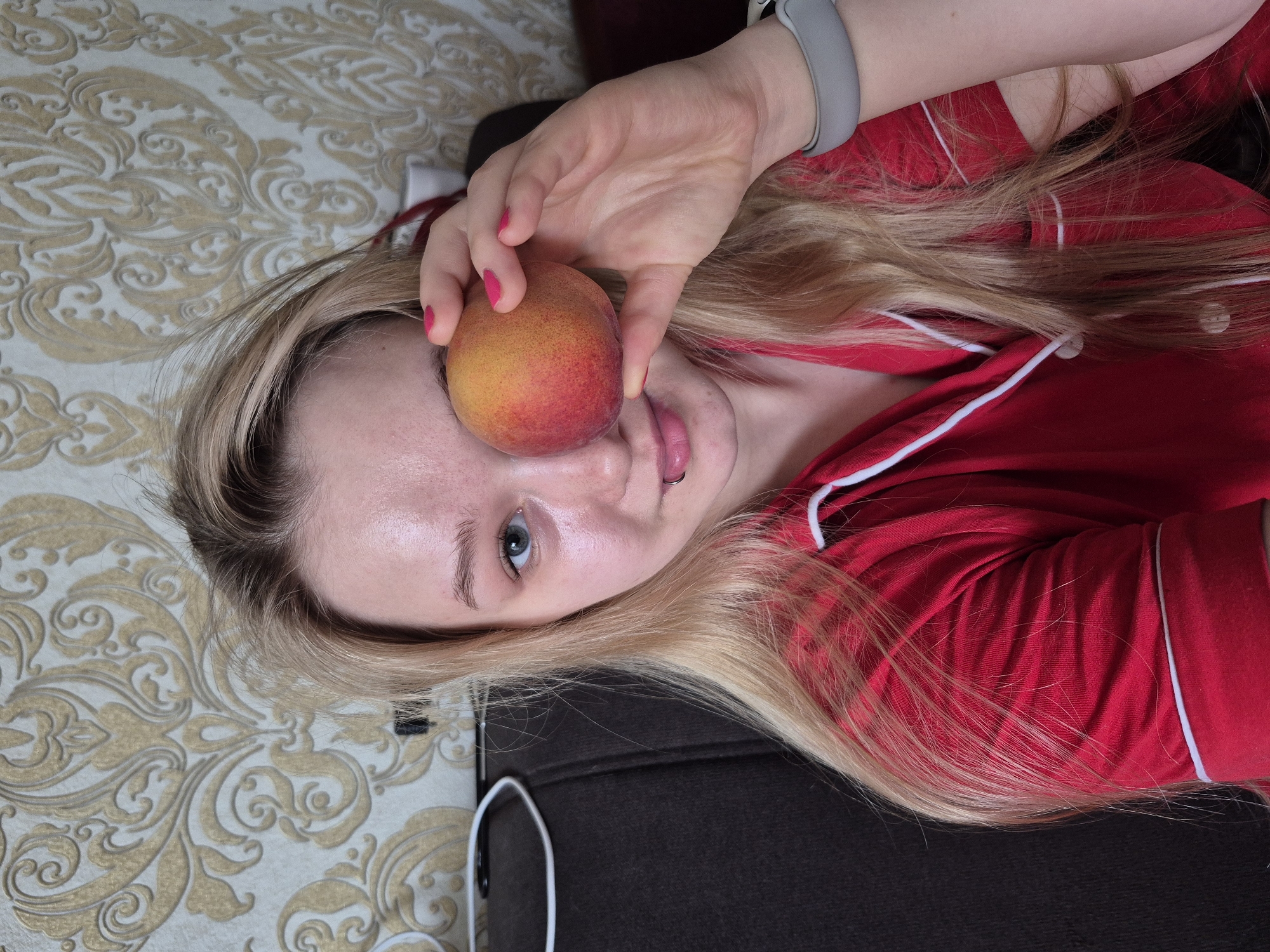 The image presents a close-up, high-angle view of a person, likely female, posing playfully in an indoor setting in Chaikovsky, Russia. The subject has long, light blonde hair that spreads across the frame and wears a red top with white piping. A grey wristband is visible on their left wrist.
With a hand adorned with bright pink nail polish, the person holds a vibrant, multi-colored peach (red, orange, and yellow hues) directly over their right eye. Their left blue eye is open and looks towards the camera. The mouth is slightly agape, revealing a lip piercing on the lower lip. The background consists of a light-colored fabric with an ornate gold and white pattern, suggesting a bedspread or similar furnishing, next to a darker, plain fabric section, possibly a duvet or sofa. The overall impression is a casual, domestic scene. No text is visible in the image.