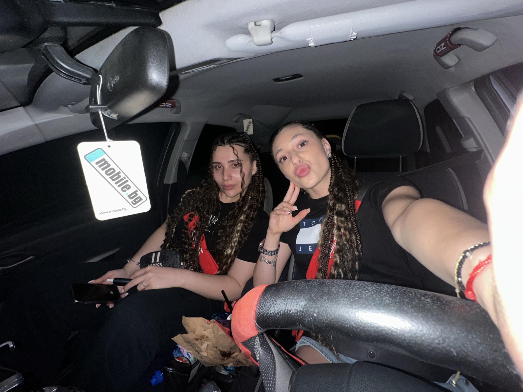 The image shows two young women sitting in the front seats of a car. The woman on the left has long, dark braided hair and is wearing a black t-shirt. The woman on the right also has long, dark braided hair and is wearing a black t-shirt with a "Tommy Jeans" logo. She is holding a phone and making a peace sign with her fingers. The interior of the car is visible, including the steering wheel, dashboard, and a rearview mirror with an air freshener hanging from it. The air freshener has "mobile.bg" printed on it. The scene appears to be taking place at night, as indicated by the dark environment outside the car. There are also some brown paper bags and other items scattered in the car. There is no discernible information in the image to indicate the location is Gabrovo, Bulgaria.