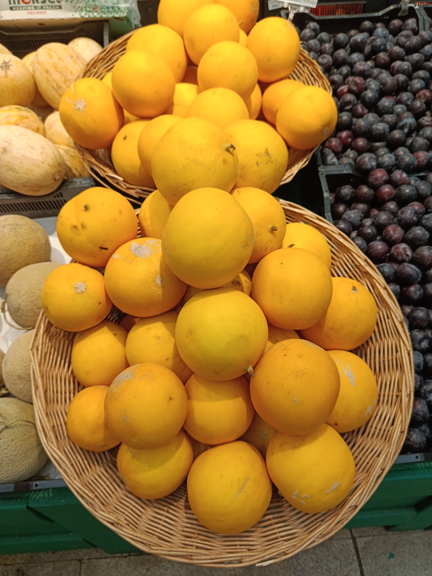 The image displays a retail fruit arrangement, likely within a grocery store or market in Moscow, Russia. The central focus is a large collection of small, round, bright yellow melons, some exhibiting minor surface imperfections, mounded within a substantial light brown wicker basket. A second, smaller wicker basket, positioned above and behind the primary one, contains additional identical yellow melons. To the right, a significant quantity of dark purple grapes is presented in dark plastic crates. A white plastic bag in the upper left background with green text confirms "TABLE GRAPES." To the left of the yellow melons, several lighter-skinned, oval melons with a distinct netted texture are visible, arranged on what appears to be a black display surface and within a black crate. The lower right portion of the image reveals a light-colored tiled floor, and a green plastic crate is partially visible in the lower left. No individuals are present in the scene.