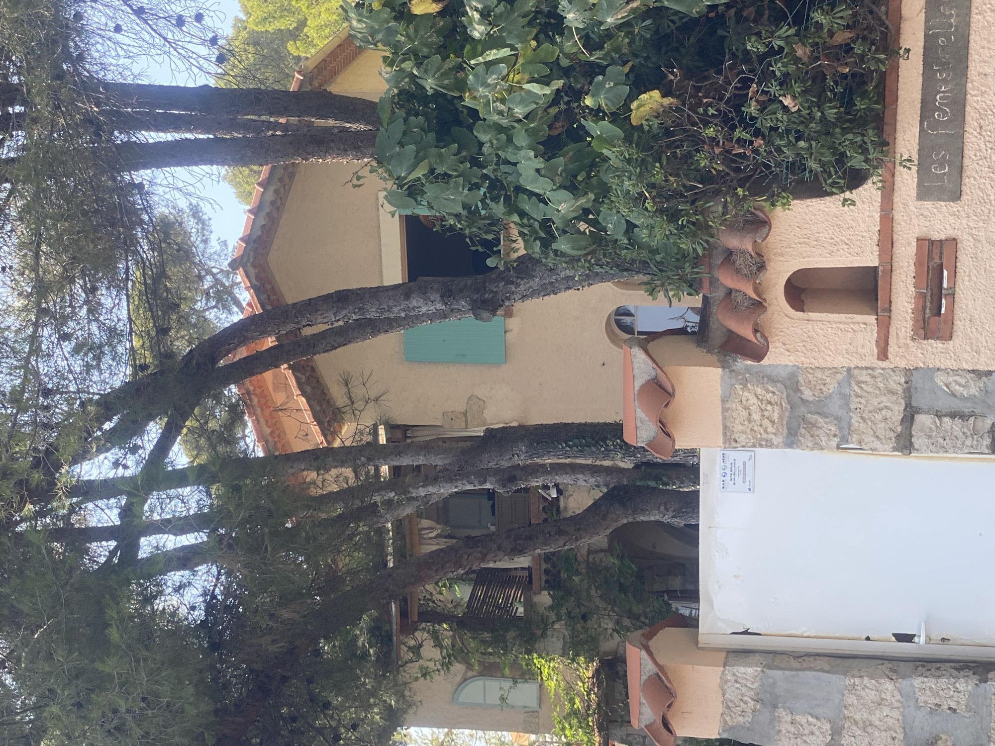 This image depicts the exterior of a building in Antibes, France, likely a residential property, surrounded by mature trees. The building has a stucco facade, a terracotta tile roof, and a balcony with wooden railings. A prominent turquoise shutter is visible on one of the windows. In the foreground, a white gate stands between two stone pillars adorned with decorative terracotta tiles. A sign on the gate reads "SAS AGIS SITE SOUS SURVEILLANCE". To the right of the gate, on a lower section of the wall, a sign in chalk-like script reads "Les Fenestrelle". The surrounding vegetation is lush and overgrown, with large trees and shrubs framing the structure. The sky is visible through the branches of the trees, suggesting it is daytime.