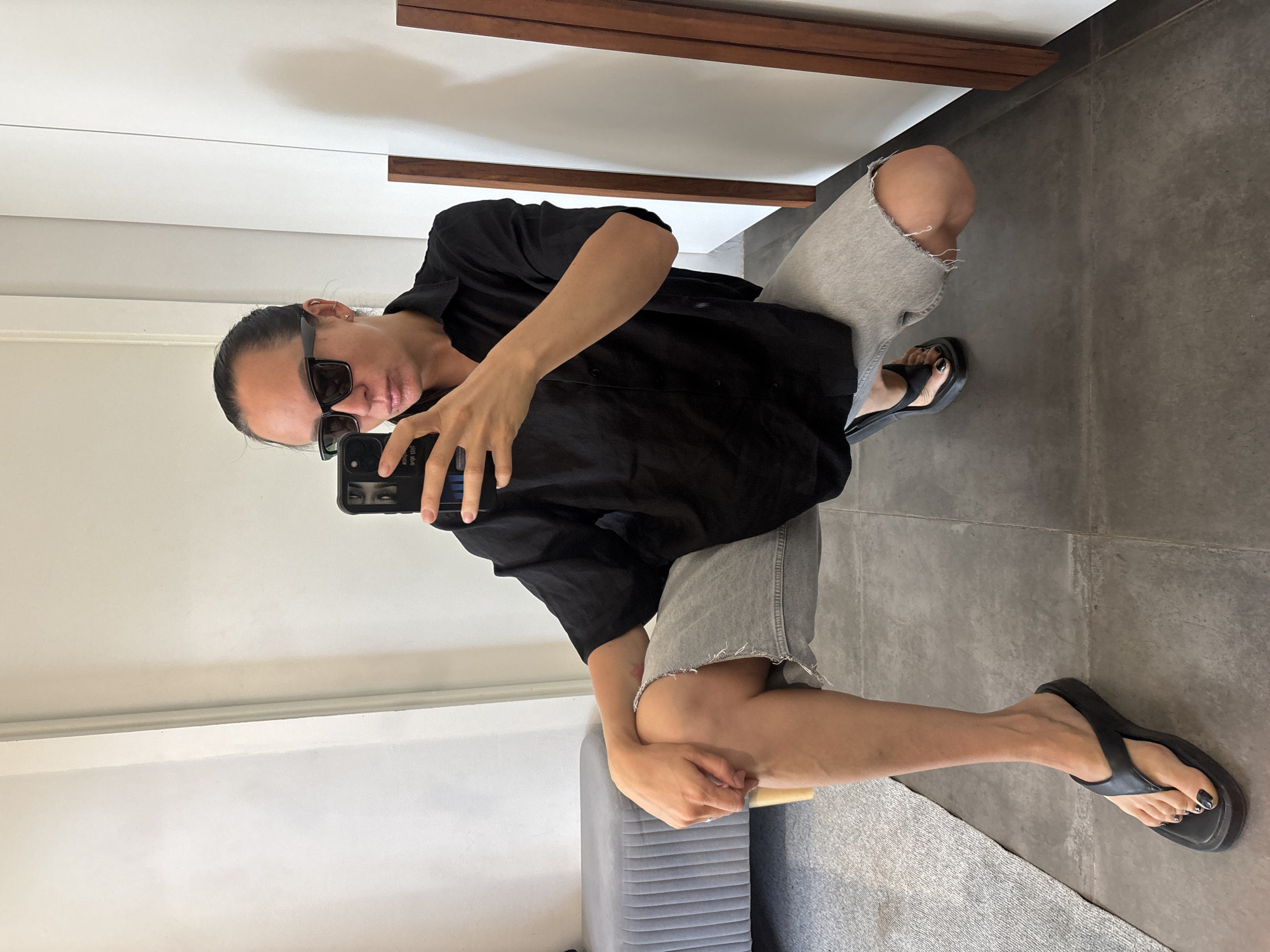 A person wearing sunglasses, a black shirt, grey denim shorts and black flip flops is crouching in a room with white walls and a grey tile floor. The person is holding a smartphone, apparently taking a selfie. The person is sitting on a small grey upholstered ottoman. There is a dark brown wooden accent on the white wall near the ceiling. The location is a room, but not specifically identifiable as being in the city of Chaikovsky, Russia.