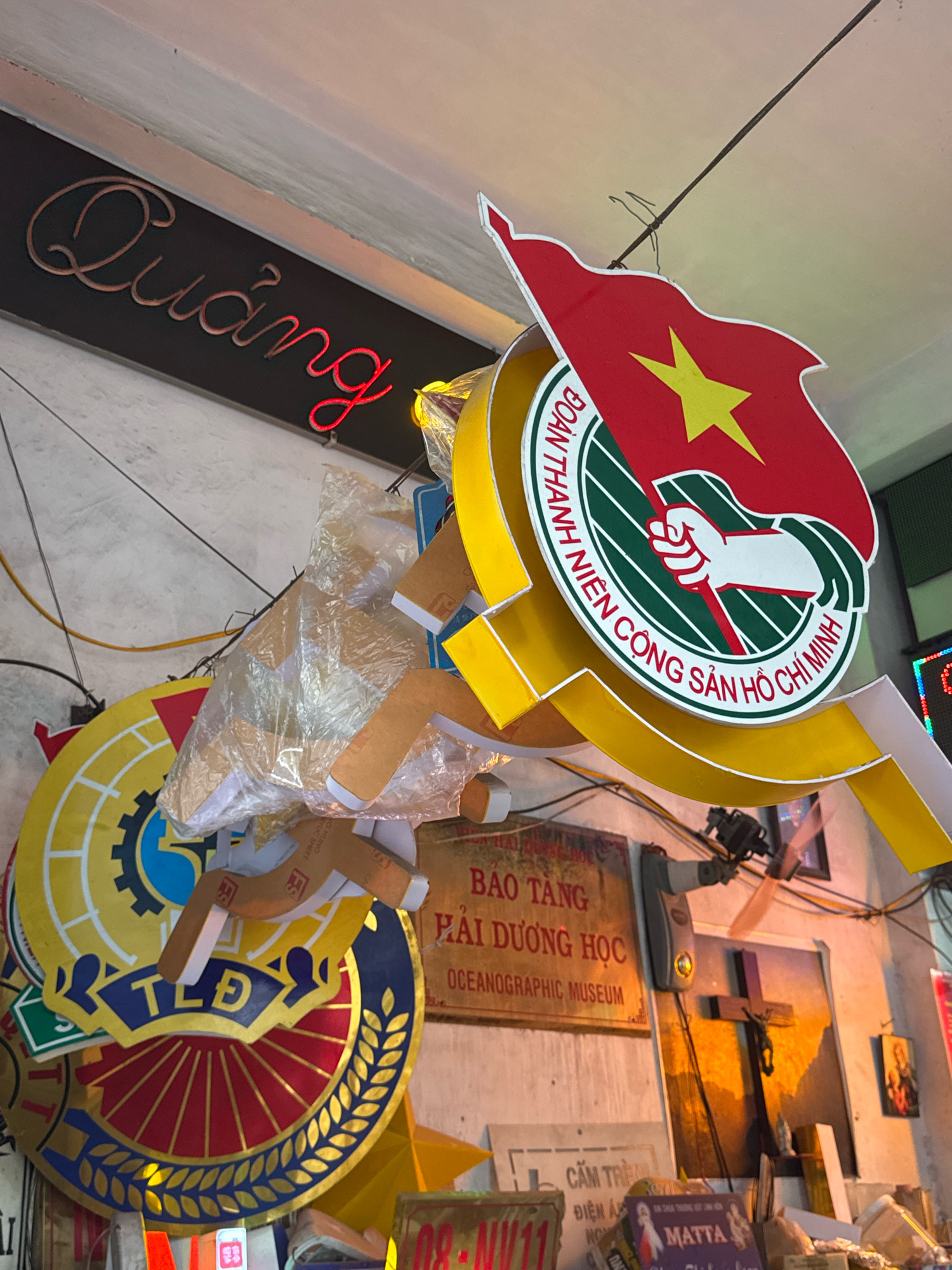 The image shows a collection of signs and objects hanging from the ceiling and walls of a shop in Nha Trang, Vietnam. In the foreground, a red and gold Vietnamese national emblem, featuring a star and a hand holding a flag, is prominently displayed. Below it, a circular sign with a yellow border and red spokes is partially visible, bearing the blue letters "TLĐ". A sign with Vietnamese text, "ĐOÀN THANH NIÊN CỘNG SẢN HỒ CHÍ MINH," is written in a circular pattern on the emblem. A neon sign with red cursive writing, "Quảng", is mounted on a dark background above the other objects.
Further back, a brass plaque reads "BẢO TÀNG HẢI DƯƠNG HỌC OCEANOGRAPHIC MUSEUM". A small electric fan is attached to the wall, pointing downwards. On the wall to the right, a framed image depicts a crucifix with Jesus. Several other signs and miscellaneous items are stacked and hanging in the cluttered environment. The lighting suggests an indoor setting, possibly a workshop or a retail space selling signs and emblems.
