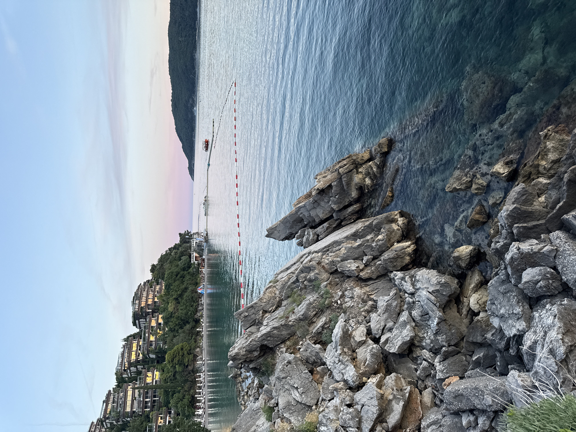 The image depicts a scenic view of a coastal area at dusk, likely located near Budva, Montenegro. In the foreground, a rocky shoreline juts into the clear, turquoise water. The rocks are varied in size and texture, with some sharp, jagged formations and smaller, more rounded stones. Green vegetation sprouts from some of the crevices in the rocks.
The middle ground features a tranquil bay with a calm sea. A floating barrier marked with red and white buoys delineates a swimming area. Beyond this, a small, red boat is visible on the water, and further in the distance, a larger vessel appears on the horizon.
Dominating the background on the left is a hillside covered with lush green trees and a cluster of modern, tiered buildings that appear to be hotels or residential complexes. These buildings are built into the slope, with balconies and windows reflecting the soft, twilight sky. The buildings are illuminated from within, suggesting it is early evening.
The sky above is a gradient of soft blues and pinks, indicating either sunrise or sunset. A distant, tree-covered island or peninsula is visible on the right horizon. The overall atmosphere is peaceful and serene. There are no people clearly visible in the immediate foreground or on the rocky shore. A few beach chairs and umbrellas are lined up on the small beach nestled between the rocky shore and the buildings.