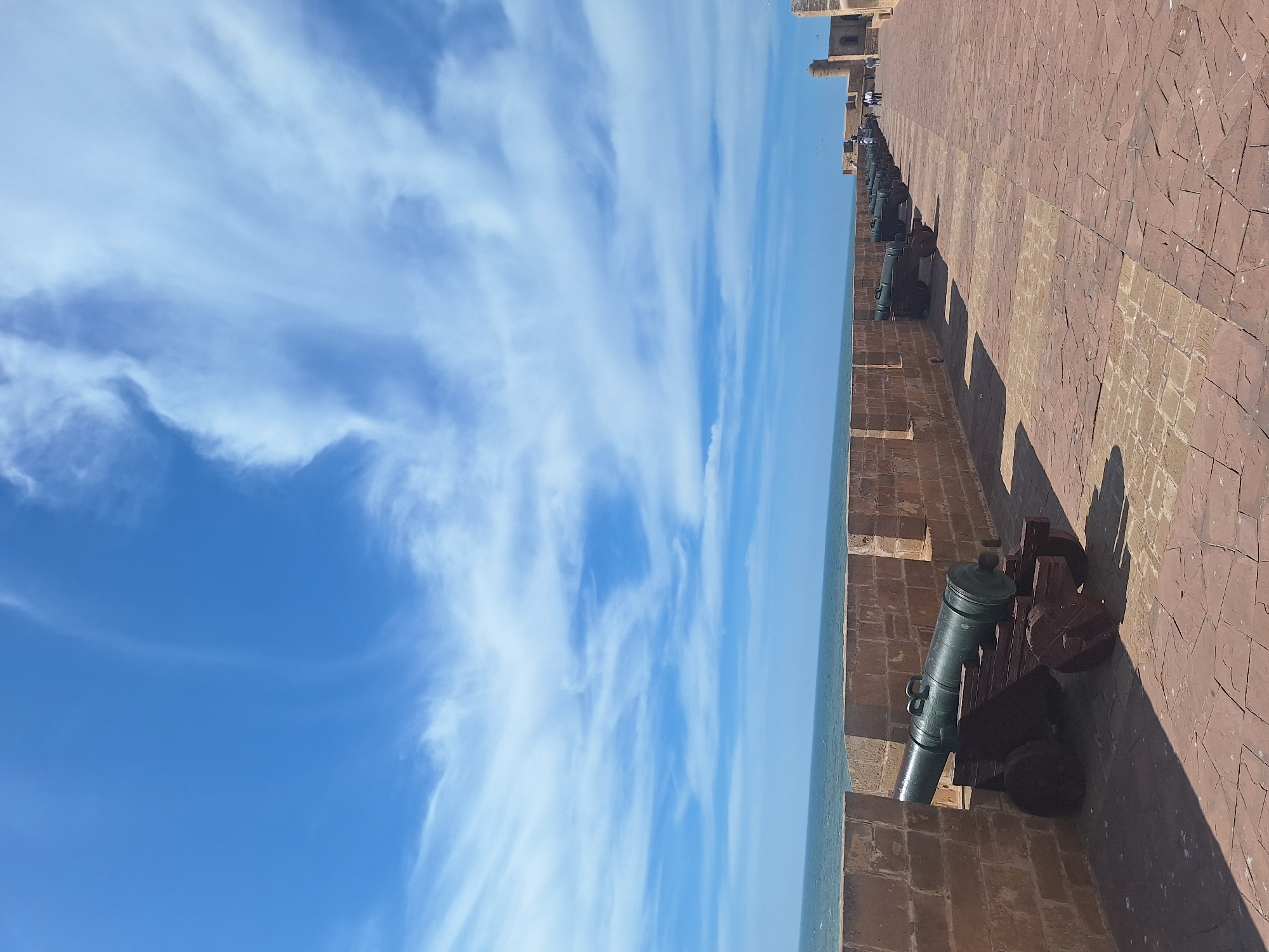 The image shows a view of the ramparts of Essaouira, Morocco, with cannons lined up along the wall overlooking the Atlantic Ocean. The sky is bright blue with wispy white clouds. The stone wall is made of reddish-brown bricks and stones. Several cannons, green with some patina and mounted on wooden carriages, are positioned along the top of the wall. A paved walkway runs along the rampart, and in the distance, there are people walking and a tower-like structure. The ocean is visible on the left, with gentle waves. The overall impression is one of historical significance and a coastal defense fortification.