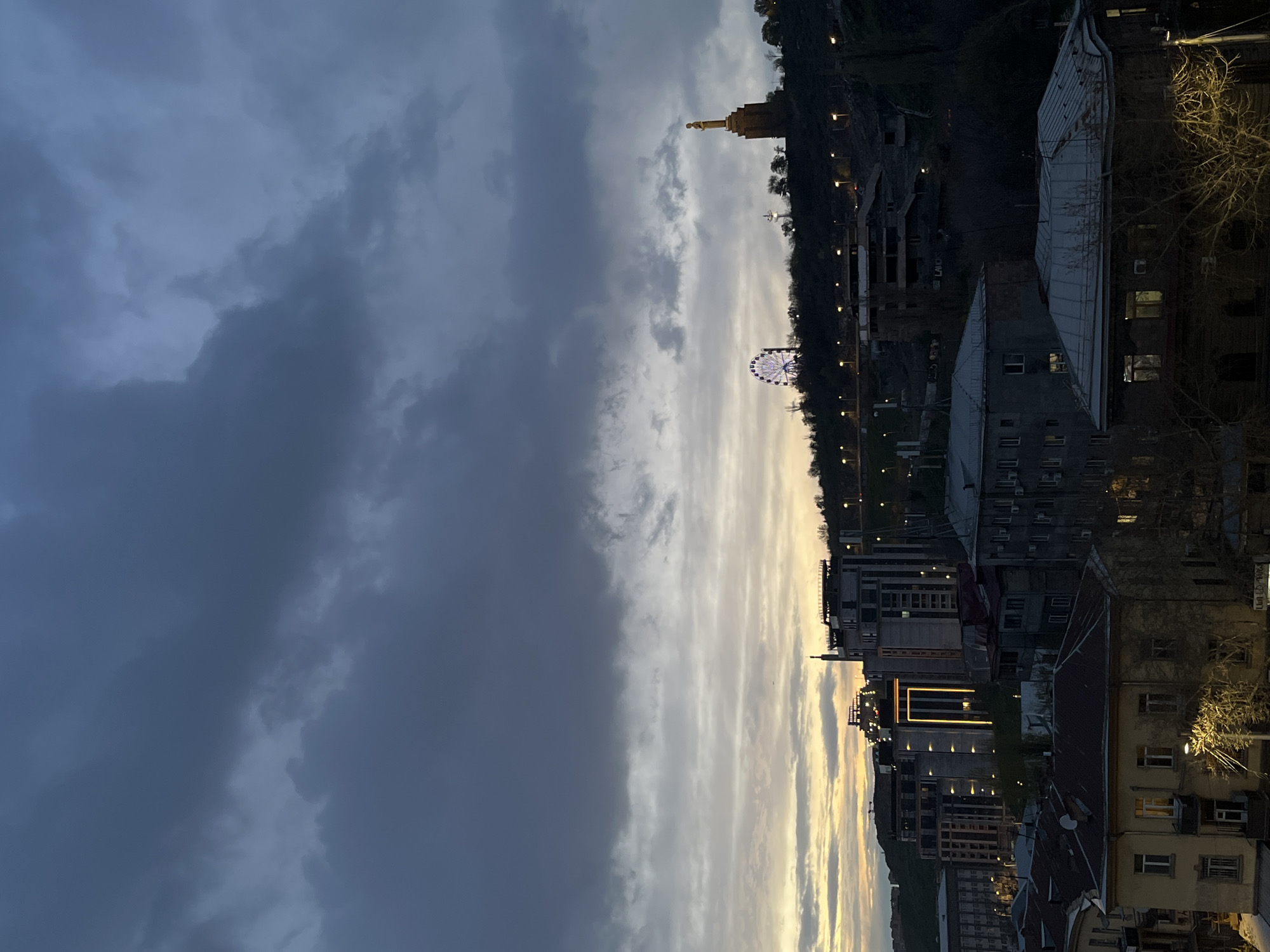 The image shows a cityscape in Yerevan, Armenia, likely taken during dusk or early evening. The sky is divided between heavy, dark storm clouds on the left and streaks of lighter, sunset-toned clouds on the right.
In the distance, a prominent golden statue atop a hill is visible, along with a Ferris wheel illuminated with white lights. A series of buildings line the lower right side of the image, varying from older, multi-story structures to more modern buildings with illuminated architectural lighting. Streetlights line the hillside, suggesting a walking path or road. The overall environment appears urban, with signs of recent construction or renovation visible in some buildings. No people are clearly visible in the image.