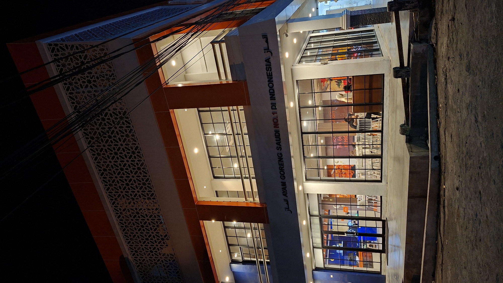 The image shows a multi-story building with an orange and white facade at night. The building has large windows, some of which reveal interior lights and what appear to be tables and chairs, suggesting it's a restaurant or cafe. There are also decorative geometric patterns on the exterior. Electrical wires are visible hanging in front of the building.
Text on the building reads "AYAM GORENG SAUDI NO.1 DI INDONESIA" and some Arabic script. The overall scene is illuminated by interior lights of the building and streetlights, creating a nighttime ambiance. The weather appears to be clear. There are no distinct people or active events clearly depicted, though reflections in the windows suggest some activity within. The location is identified as Lhokseumawe, Indonesia.