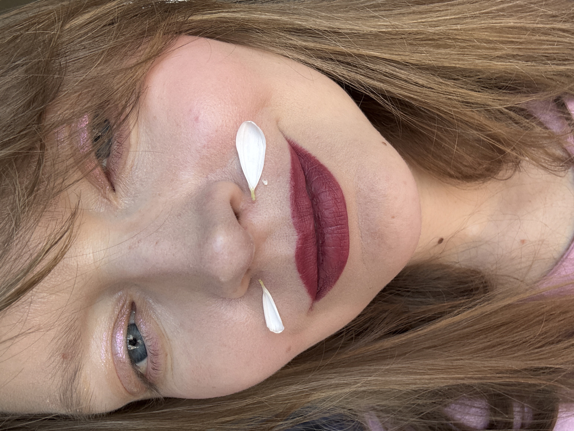 A close-up portrait shows a young woman lying on her side. Her face is tilted towards the camera, framed by long, light brown hair. Two white flower petals rest on her face, one on her nose and another near her mouth. She has fair skin and light blue eyes, with a hint of pink eyeshadow. Her lips are painted with a deep red matte lipstick. The image does not appear to have been taken in Izhevsk, Russia.