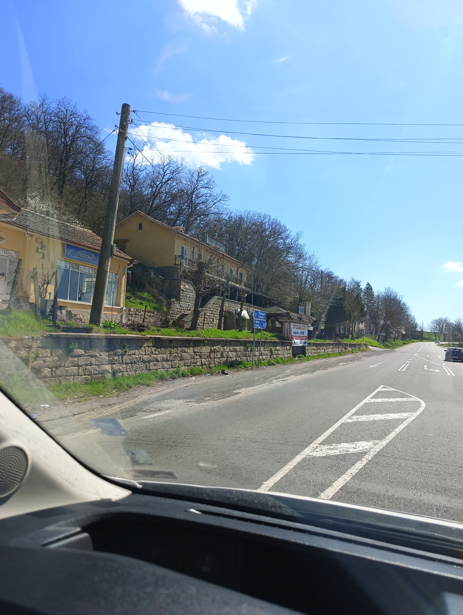 The image depicts a view from inside a vehicle on a road outside the city of Unknown, Unknown. The scene captures a two-lane asphalt road with white painted lane markings, including chevron patterns. On the left side of the road, there is a retaining wall topped with greenery, behind which various buildings are situated on a gentle slope. A utility pole is visible on the left side of the image, with power lines stretching across the sky. The sky is blue with scattered clouds. A car is visible further down the road, in the right lane. The foreground shows the dashboard and part of the windshield of the vehicle from which the image was taken.