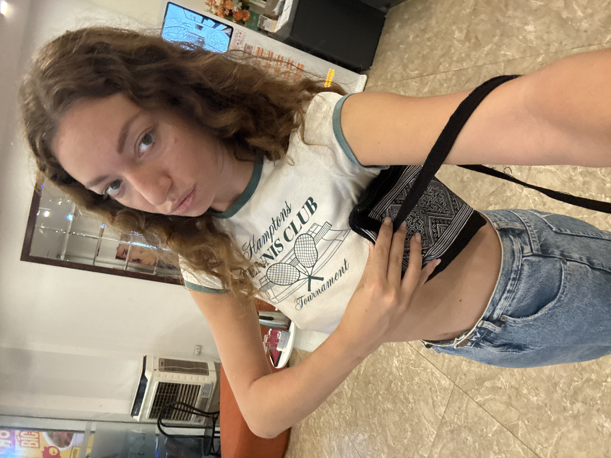 A young woman with curly brown hair takes a selfie in a room with a tiled floor. She is wearing a white t-shirt with a green trim and a graphic that reads "HAMPTONS TENNIS CLUB Tournament" with two tennis rackets. She is also wearing blue jeans. A black bag with a patterned strap is slung over her shoulder and she is holding the strap with her right hand. In the background, a wall-mounted air conditioner is visible. A digital display screen is mounted on the wall and shows an image. The lighting suggests it is daytime.