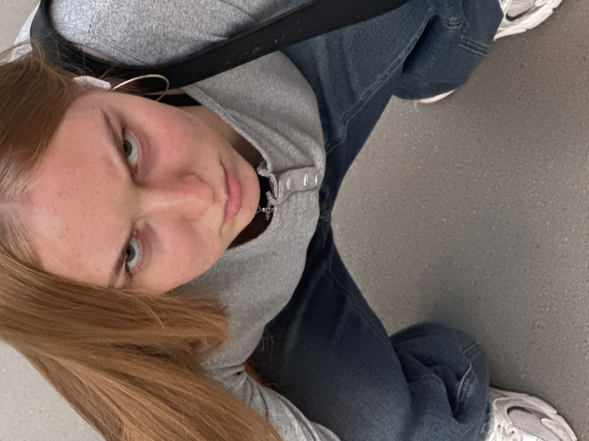 An individual, appearing to be a young adult female, is depicted crouching outdoors in an urban environment. She has long, straight, reddish-brown hair and fair skin. Her facial expression is stern and direct, with furrowed brows, narrowed light-colored eyes, and a slightly pursed mouth, indicating an intense gaze aimed at the camera. She wears a light grey long-sleeved Henley shirt with visible buttons at the neckline, loose-fitting blue denim jeans, and white and grey athletic shoes. A white wireless earbud is visible in her right ear, along with a silver-colored hoop earring. A silver-colored pendant hangs from a necklace around her neck. A black strap, possibly from a bag or backpack, is visible over her left shoulder. The ground beneath her is a light grey, textured paved surface, consistent with concrete or asphalt. The background is out of focus, showing a light-colored, indistinct surface typical of an urban setting. The location is an urban area, consistent with the city of Perm, Russia.