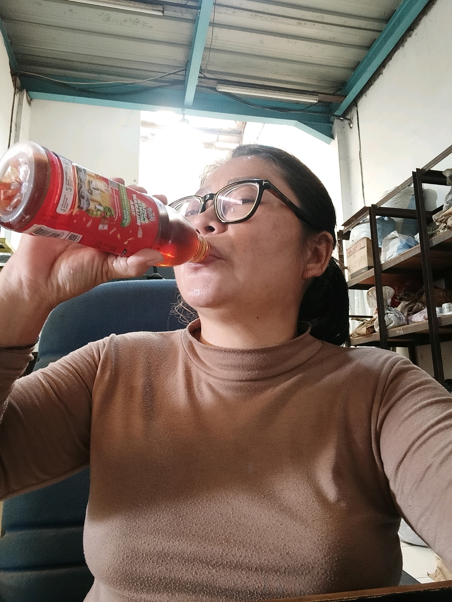 A woman with dark hair and glasses is drinking from a red bottle of liquid. She is wearing a brown, long-sleeved shirt. The background shows a corrugated metal ceiling and white walls, with shelves filled with items. The scene appears to be indoors, possibly in a workshop or storage area, and is located in Belimbing, Indonesia.