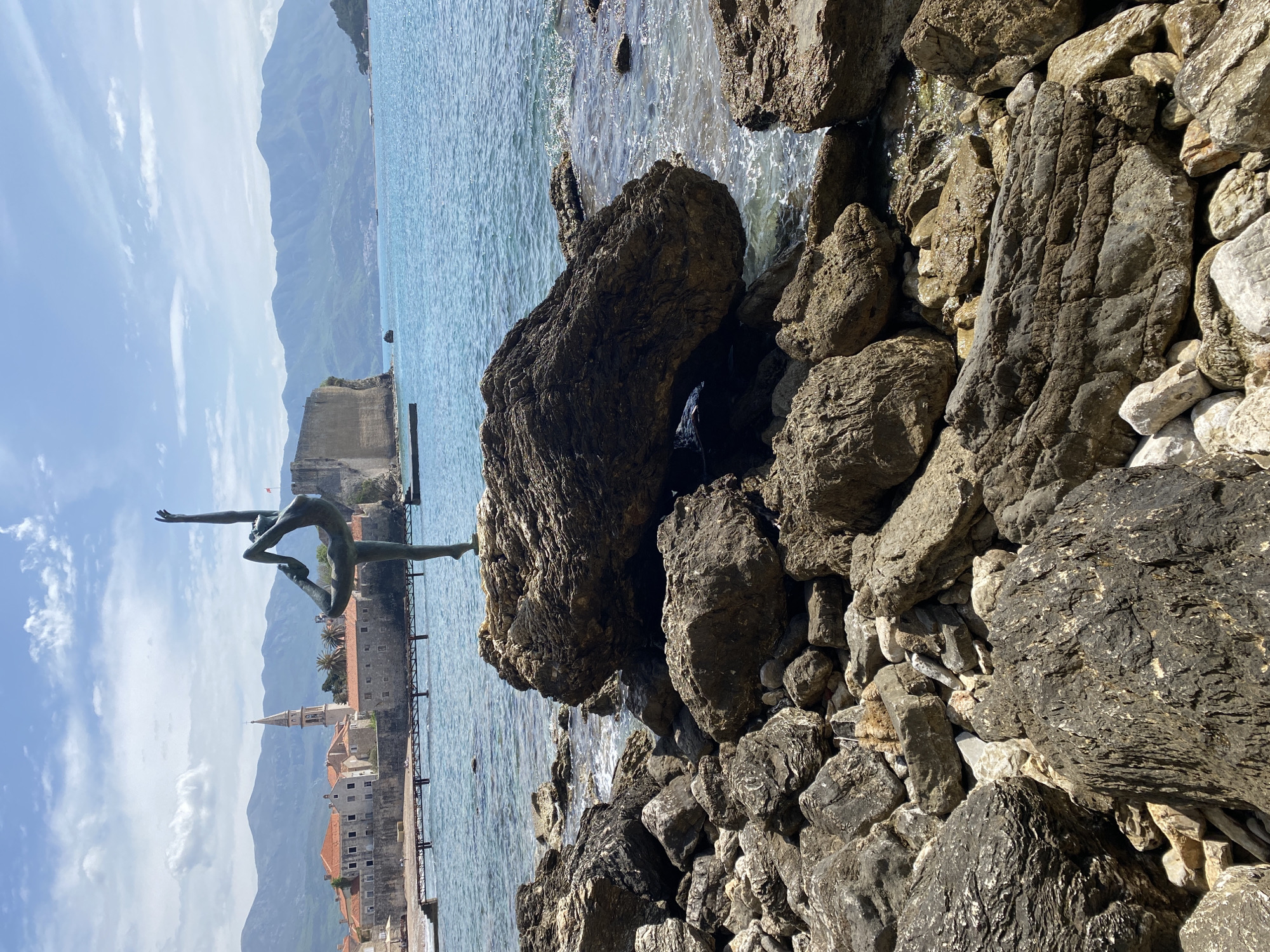 The image depicts a bronze statue of a gymnast balanced on a rock by the sea in Budva, Montenegro. The statue is positioned against a backdrop of the old town, with its medieval walls, buildings, and a church steeple visible. In the background, mountains rise in the distance. The sea is a clear blue, and large rocks and stones are situated in the foreground along the shore. The sky is partially cloudy. The photo is taken at a slight angle, with the statue in the left part of the image.