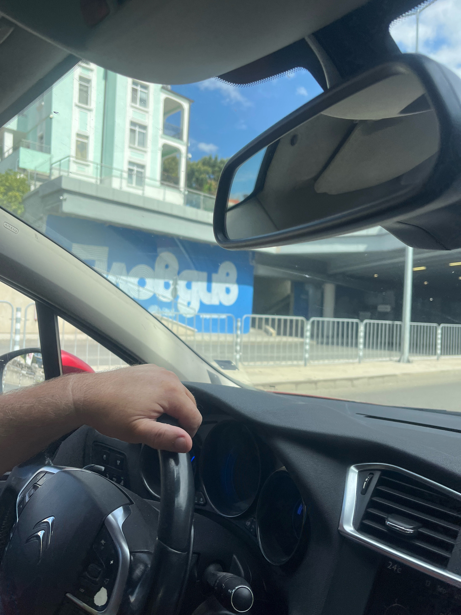 The image is a first-person view from inside a car in Plovdiv, Bulgaria, with a hand on the steering wheel. The driver's side of the car's interior is visible, including the dashboard with air vents and part of the steering wheel with a Citroën logo. Outside the car, through the windshield and side windows, a light blue building with white trim and balconies is seen, along with a blue banner with white lettering that appears to read "JUMBO". This suggests the location is near a Jumbo store in Plovdiv. A metal barrier fence runs along the foreground outside the car. The sky is clear and blue with some clouds. The time of day appears to be daytime, likely late morning or afternoon, given the lighting.