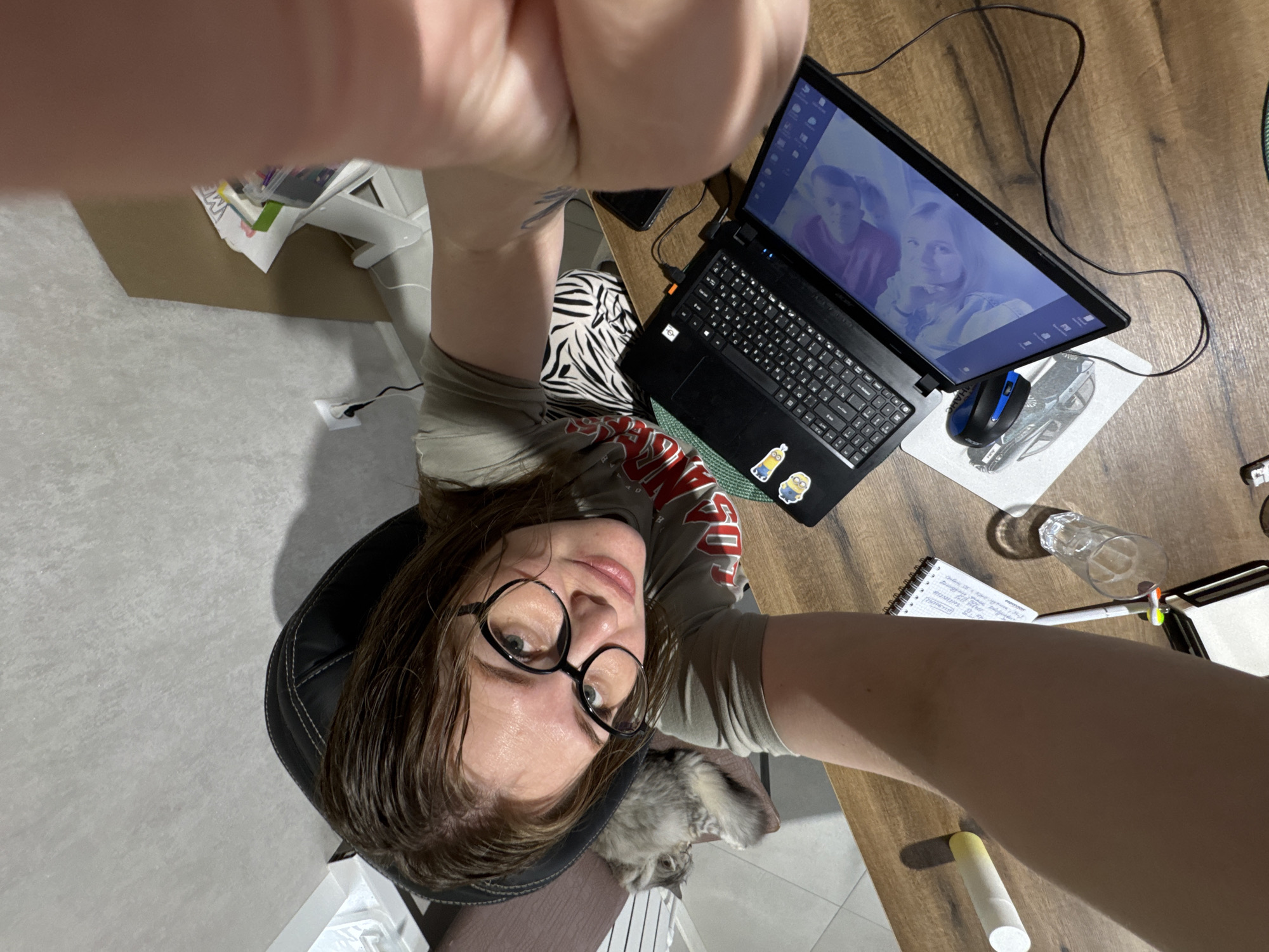 A woman wearing glasses and a t-shirt is taking a selfie from an overhead angle. She is seated in a black office chair with a gray cat lying near her. A laptop is on a wooden desk next to her. The laptop screen displays a video or picture of two people. A mouse, mousepad, notebook, pen, and a glass of water are also on the desk. Part of the desk is covered in what seems to be a patterned fabric. The background includes the floor and a white storage unit. The location is possibly in a home or office. There is no evidence of being in the city of Gornyi (Dvurechenskoe S/P), Russia.