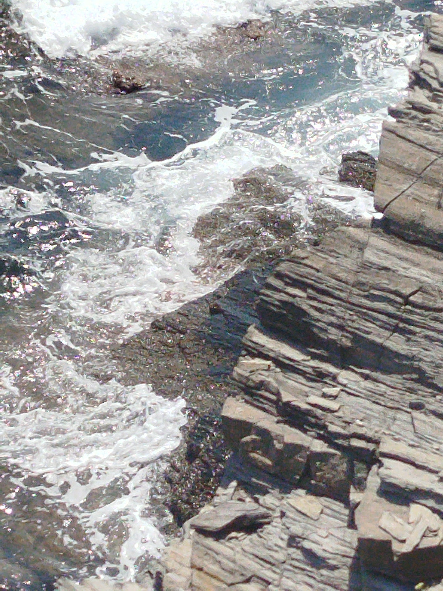 A rocky shoreline in Sithonia Municipal Unit, Greece, is captured in an overhead view. The water is a mix of deep gray and shimmering turquoise, with white waves crashing against dark, wet rocks. The rocks are layered and striated, varying in color from light gray to deep brown. The overall impression is one of natural ruggedness and the dynamic interplay of water and stone. There are no people or man-made structures visible.