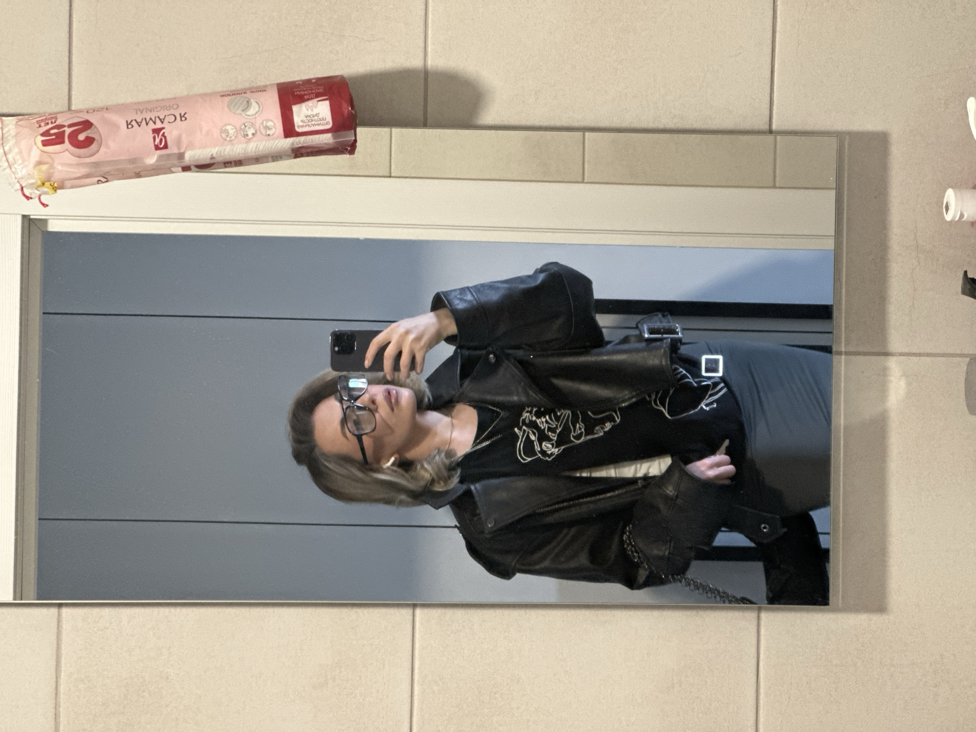 A person with blonde hair and glasses is taking a mirror selfie in what appears to be a bathroom. The person is wearing a black leather jacket and a black t-shirt with a white design. On the wall above the mirror, there is a package of cotton pads. The location of Khimki, Russia is inferred from the Cyrillic text on the package. There is a toilet paper holder to the right of the mirror. The bathroom has light-colored tile walls.