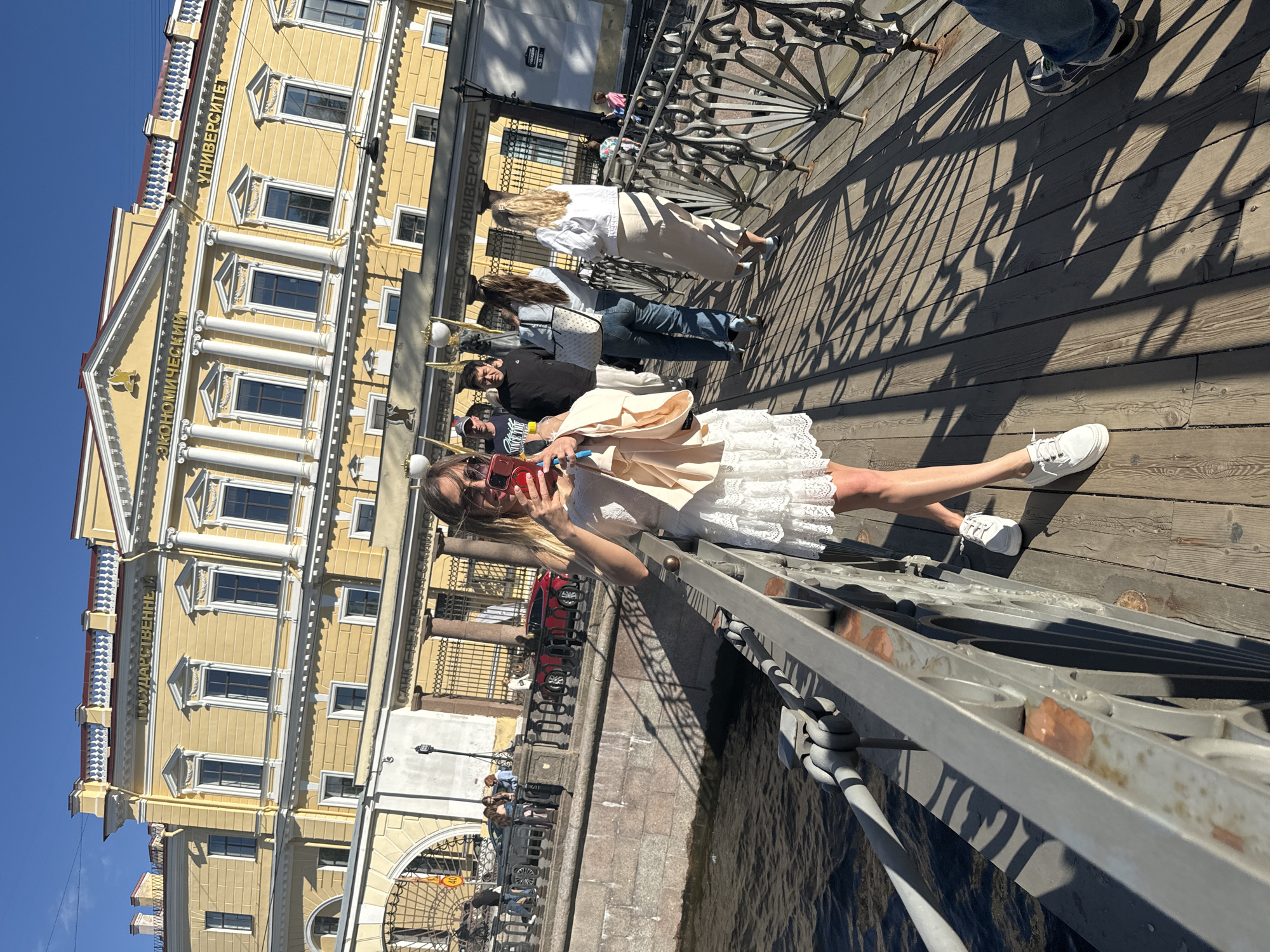 The image depicts a sunny day in Saint Petersburg, Russia, likely during the daytime. The scene is set on a bridge overlooking a canal, with a grand, yellow neoclassical building as a backdrop. The building features architectural elements like columns, arched windows, and decorative cornices, and visible text indicates it is related to the "ST. PETERSBURG STATE ECONOMIC UNIVERSITY" (CANKT-ПЕТЕРБУРГСКИЙ ГОСУДАРСТВЕННЫЙ ЭКОНОМИЧЕСКИЙ УНИВЕРСИТЕТ).
Several people are on the bridge, walking and posing. In the foreground, a woman in a white lace dress is taking a selfie with her phone. She is wearing sunglasses and white sneakers. Behind her, other individuals are seen walking, some in casual attire. One person is wearing jeans and a t-shirt, while another is in a white shirt and skirt. The bridge itself has a decorative metal railing and a wooden walkway. Sunlight casts long shadows across the walkway, indicating a bright day. The water in the canal below is rippling slightly. The overall atmosphere appears relaxed and cheerful, consistent with a pleasant day out in the city.
