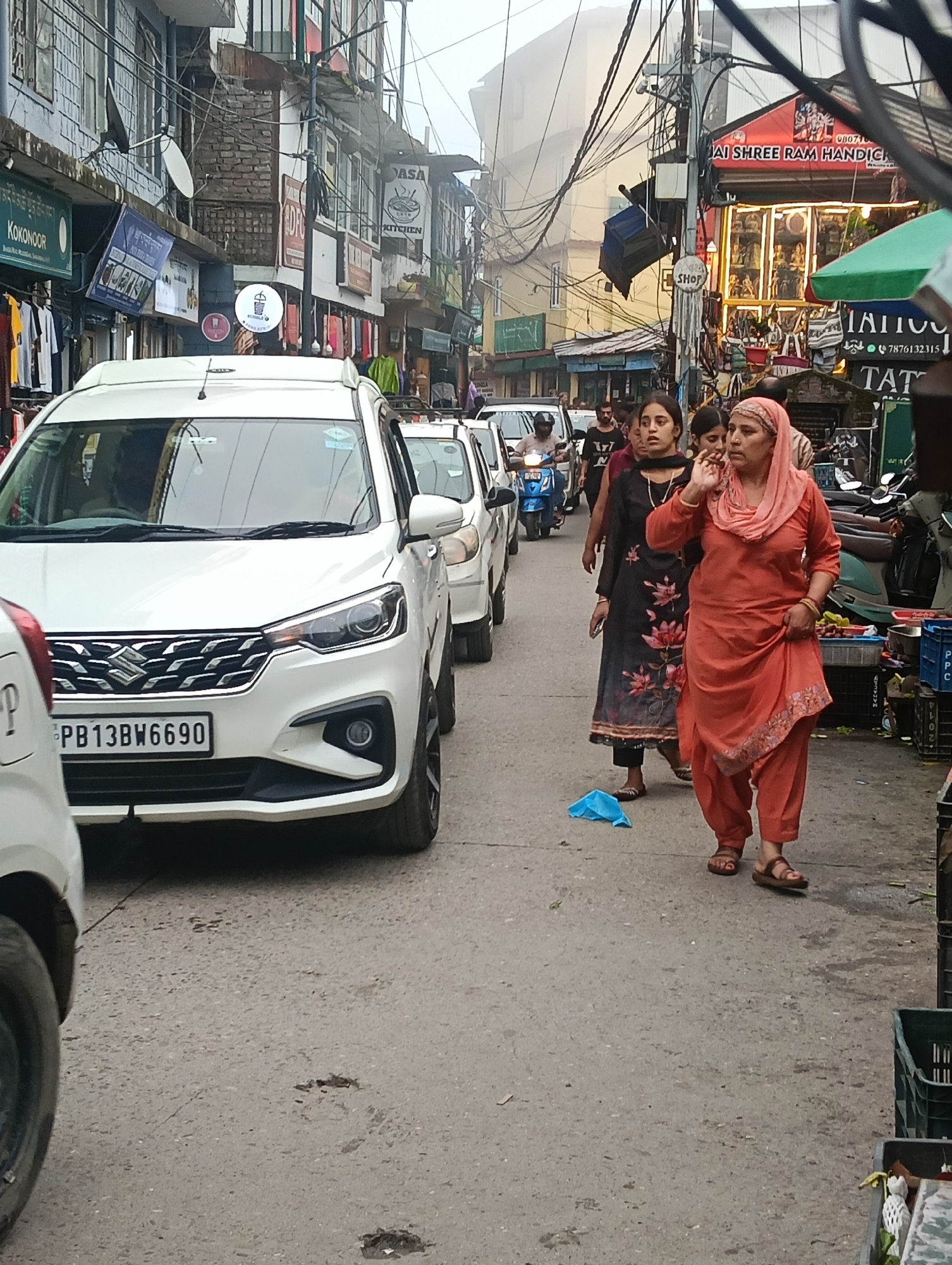 A street scene in Dharamkot, India, shows a white multi-purpose vehicle in the foreground with license plate PB13BW6690. Behind it, a white car and a blue scooter are visible. Several people are walking on the street, including a woman in an orange salwar kameez and a woman in a black floral dress. Shops line the street, displaying various goods and signage. The environment appears to be an urban marketplace with overhead wires and a slightly overcast sky.