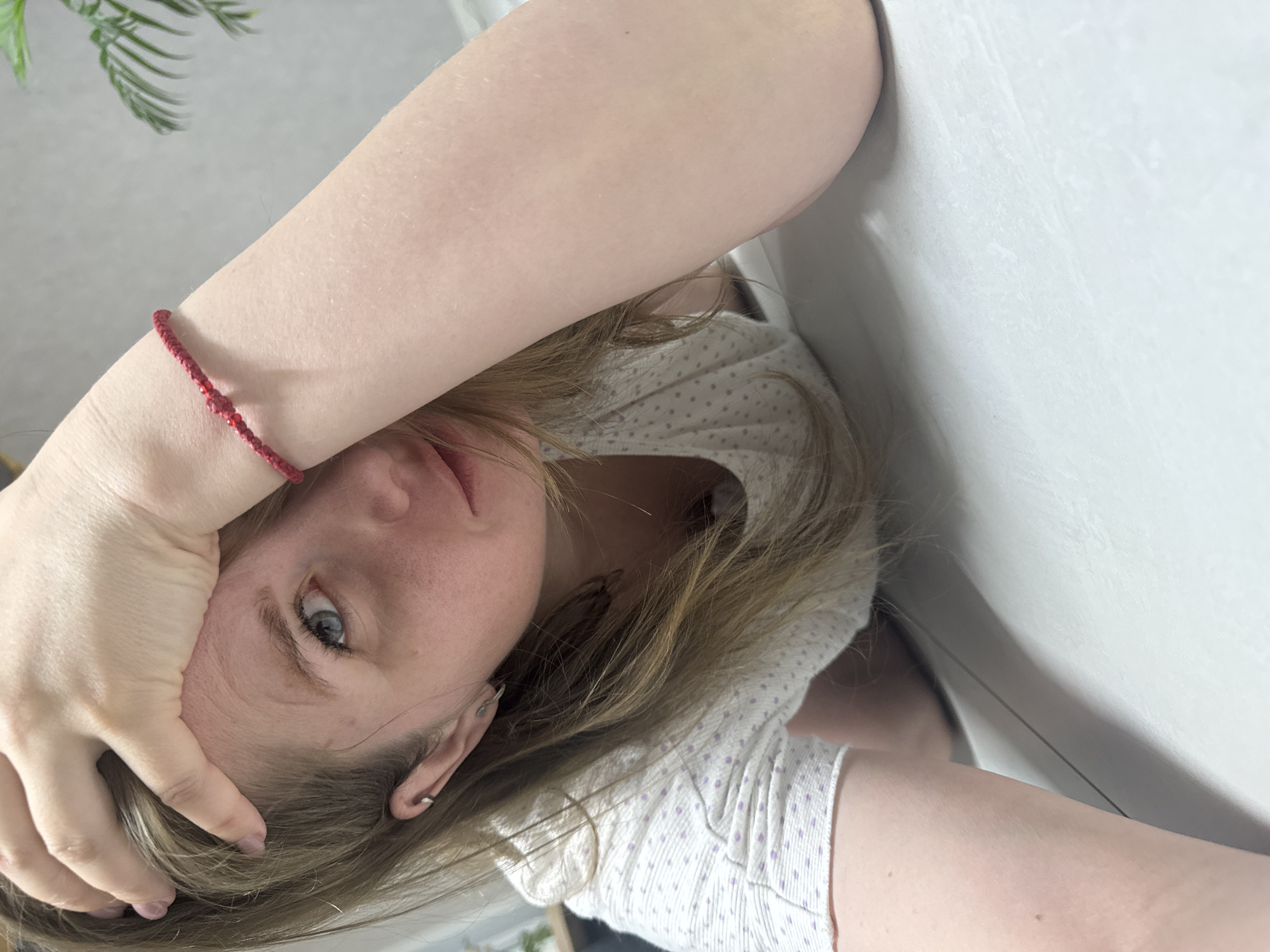 A young woman is photographed from an unusual angle, possibly lying down or tilting her head. She has fair skin, blonde hair, and blue eyes. She is wearing a white top with small purple polka dots. Her right arm is raised, with her hand resting on her forehead. A red bracelet is visible on her wrist. The background includes a white wall and a glimpse of greenery, possibly a plant. The image does not contain enough information to specifically determine if the location is Gornyi, Russia.