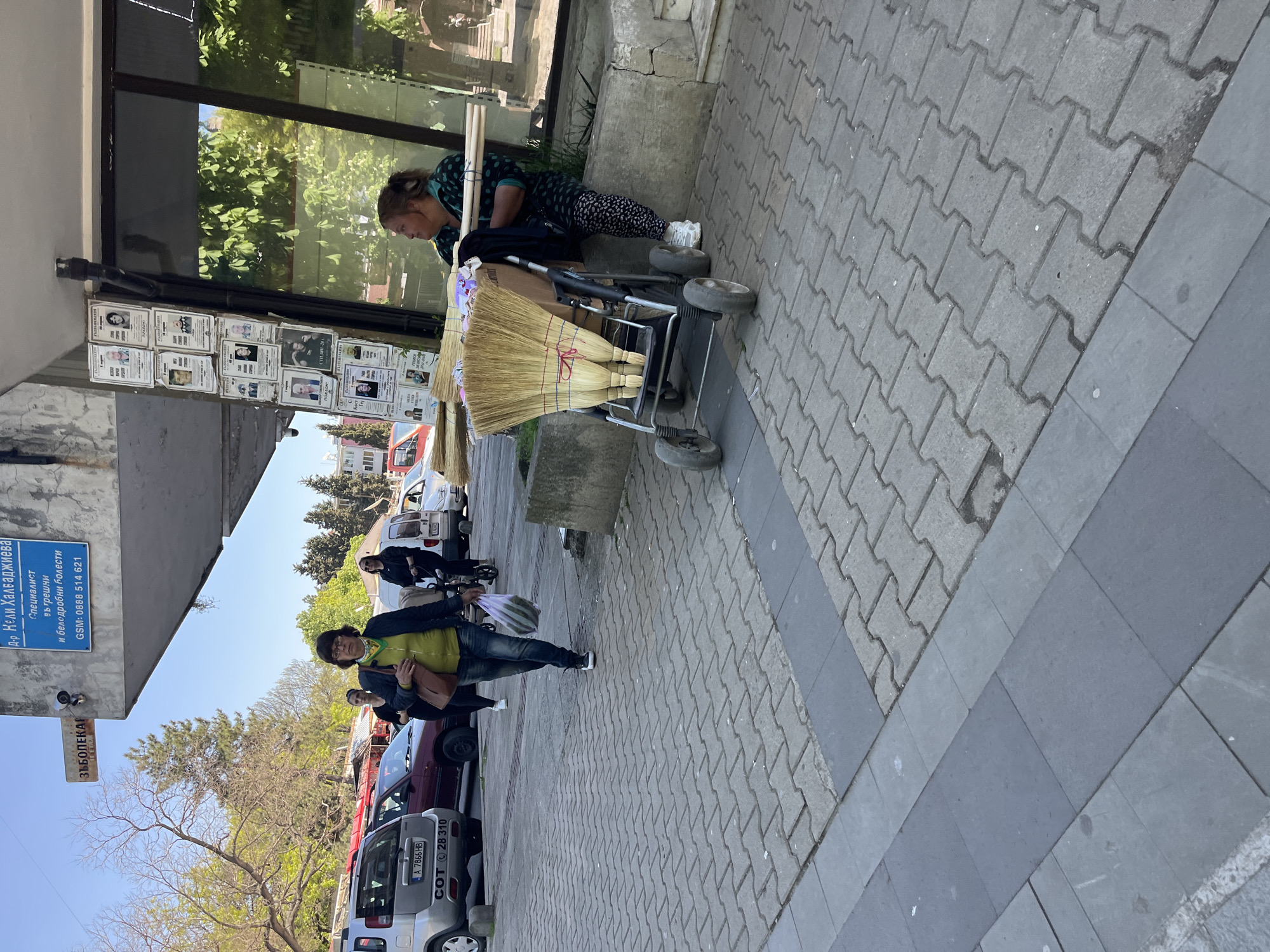 The image depicts a street scene in what appears to be Karnobat, Bulgaria. A woman is seated next to a stroller loaded with brooms, possibly for sale. Nearby, a sign indicates the location of Dr. Keli Halvadzhieva, a specialist in internal and pulmonary diseases. Two other people walk along the sidewalk. A line of parked cars is visible along the street.