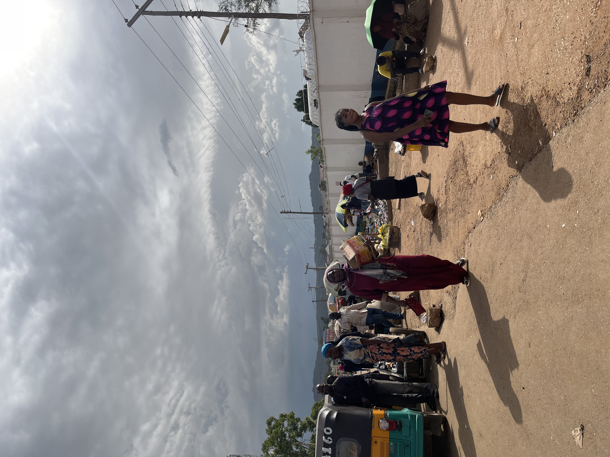In Rafin Par, Nigeria, a busy street scene unfolds under a cloudy sky. Several people are gathered along a dirt road, some standing, others sitting. A woman in a vibrant pink and purple polka-dot dress is prominently featured in the foreground, facing the camera with a slight smile. She is wearing sandals and holding a phone. To her left, another woman in a maroon dress and headscarf is also visible. In the background, more individuals are seen, along with various objects including boxes, umbrellas, and what appears to be merchandise for sale. A three-wheeled auto-rickshaw, a common mode of transport in the region, is parked on the left side of the frame. Power lines crisscross the sky, and a white wall with barbed wire adorns the top of a fence in the distance. The overall impression is of a bustling marketplace or a busy street corner, likely during daylight hours given the shadows cast by the people and objects. The weather appears overcast, with dramatic clouds contributing to the atmosphere.