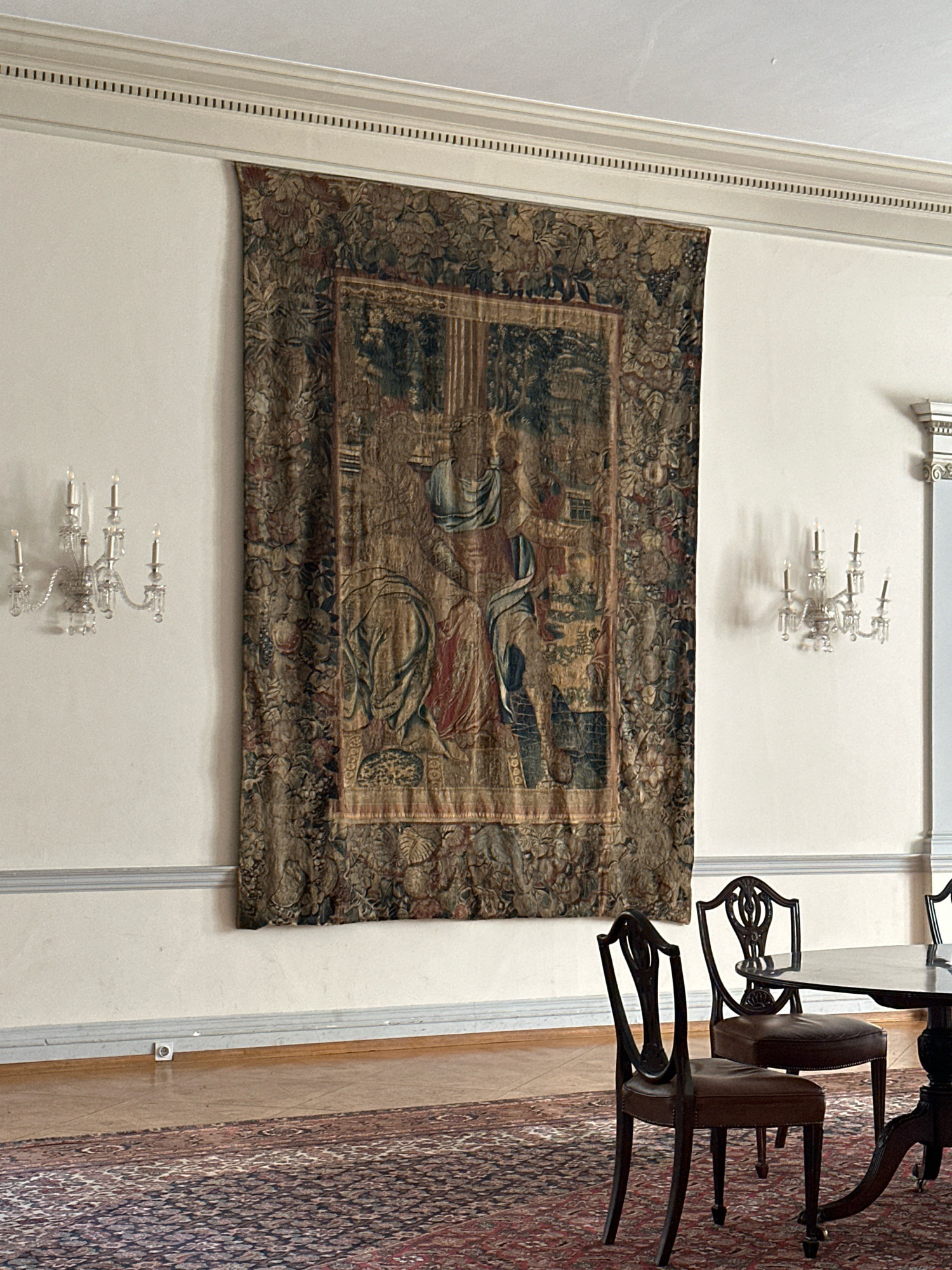 This is a photograph of an interior scene. The focal point is a large, ornate tapestry hanging on a beige wall. The tapestry depicts a scene with figures in historical attire, rendered in muted, earthy tones. Below the tapestry, a patterned rug covers the wooden floor. Two dark wooden chairs with brown leather seats are positioned near a dark, round dining table. On the wall to the left of the tapestry, a crystal sconce with multiple candle-like lights is mounted. Another similar sconce is visible on the right side of the tapestry, casting shadows on the wall. The room features white trim along the top and bottom of the walls, with a decorative molding at the ceiling. The overall impression is of an elegant, traditional interior space. The location in Belgrade, Serbia, is not discernible from the image itself.