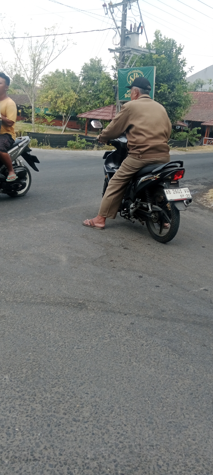 A middle-aged man wearing a light brown jacket, light brown pants, and a black baseball cap rides a black Honda motorcycle on a paved road. He is wearing brown sandals. The motorcycle has a license plate that reads "AB 2601 VU". The man is in motion, riding alongside another person on a motorcycle who is partially visible on the left side of the image. The background shows trees, buildings with red-tiled roofs, and a utility pole with electrical equipment. A green banner with "SOTO LAMONGAN" and "SPC" is visible on the utility pole. The scene appears to be in a semi-urban area in Maguwoharjo, Indonesia, likely a roadside setting. The sky is overcast.
