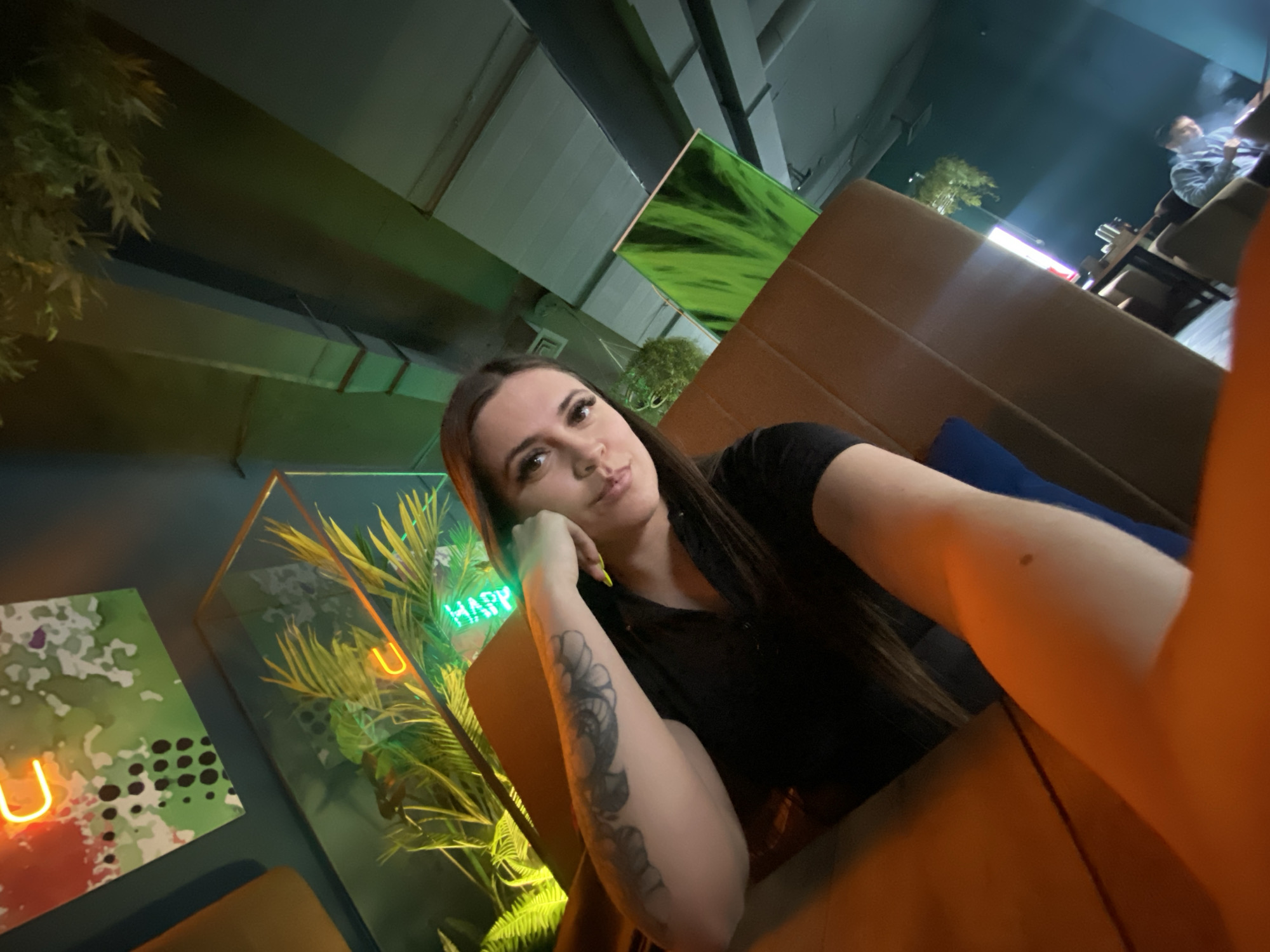 A woman with long brown hair and a tattoo on her left arm is sitting on a brown couch in what appears to be a restaurant or lounge. She is wearing a black top and resting her head on her hand. In the background, there are leafy decorations and neon signs. In the distance, there is a table with people sitting around it. The location is potentially a restaurant or lounge in the city of Chaikovsky, Russia.