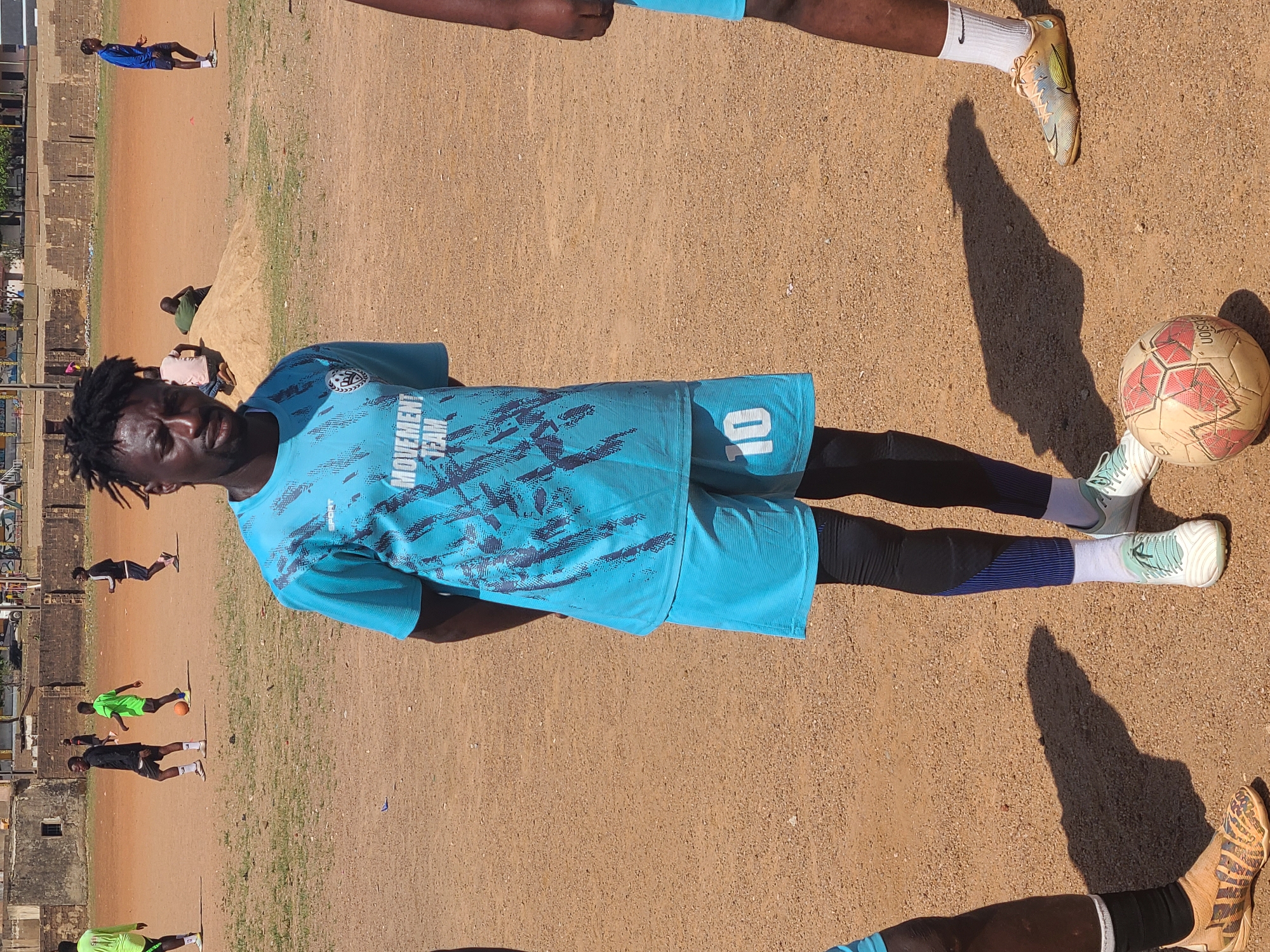 A close-up, rotated, medium shot shows a man in a blue soccer uniform, posing, on a sandy field, likely in the city of Unknown, Unknown. The man has dark skin, short dreadlocks, and is wearing a light blue soccer jersey with the words "MOVEMENT TEAM" on it, along with the number "10". He's also wearing matching blue shorts, black leggings, and white socks with light green sneakers. A worn soccer ball sits next to his feet. In the background, other people are seen playing soccer on the same sandy field, with a building visible in the distance. A leg of another person wearing soccer shoes is visible.