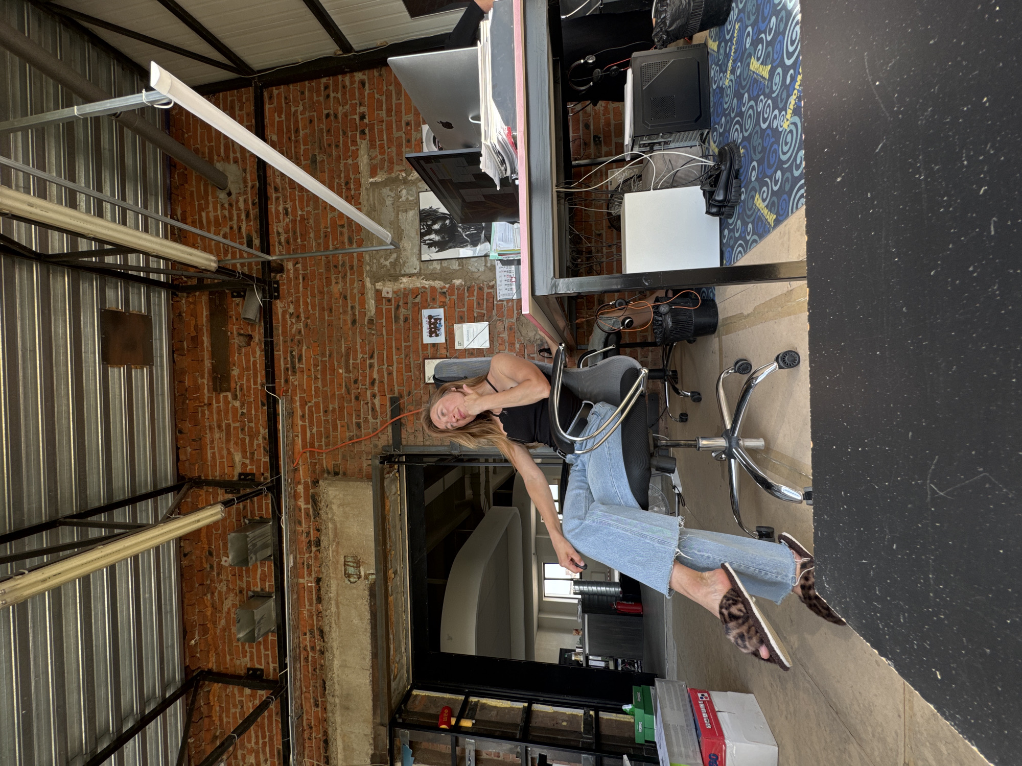 A woman is sitting on a chair in what appears to be an industrial or renovated space. The location is difficult to determine, but if specified to be in Chaikovsky, Russia, the description focuses on the visual elements present.
The woman has light brown hair and is wearing a black tank top and blue jeans. She is sitting in a swivel chair with a chrome base. Her feet are clad in animal-print slippers.
Behind her is a brick wall and an open doorway or archway. To the left, the ceiling has exposed metal beams and fluorescent lighting.
To her right, there's a desk with computer equipment, including a monitor, keyboard, and a desktop tower. A patterned rug is under the desk.
The room appears to be in a building that may have been converted for office or residential use, as evidenced by the mix of industrial elements and office furnishings.