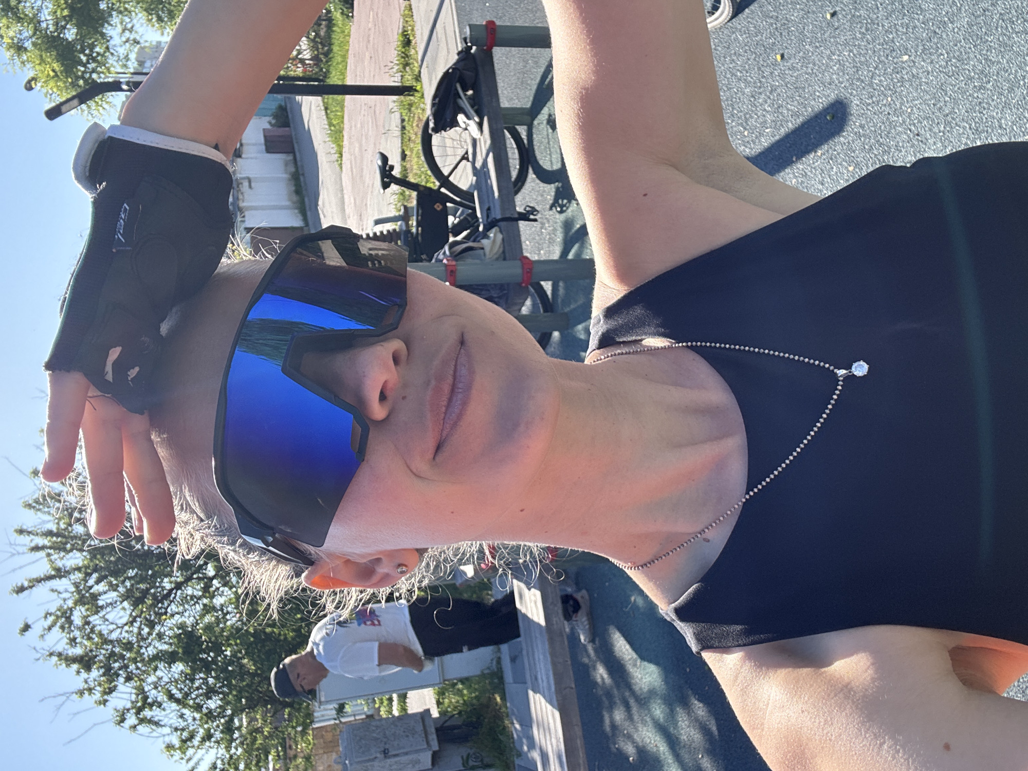 A close-up, rotated view of a person wearing sunglasses, a black tank top, and a cycling glove. A bicycle and a person in a white shirt are visible in the background. The location is presumed to be Мысовское сельское поселение, Ukraine, based on the user's prompt.