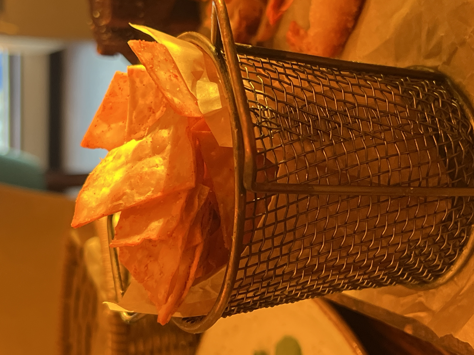 A close-up, eye-level shot focuses on a small, wire mesh basket filled with golden-brown tortilla chips. The chips are layered and appear to be seasoned, with hints of red spice visible on their surfaces. White parchment paper lines the bottom of the basket, peeking out from between the chips. The basket itself has a metallic sheen and a handle on the right side. The background is softly blurred, suggesting an indoor setting with warm, ambient lighting. To the right of the basket, out of focus, are more food items, possibly fried shrimp. The overall scene evokes a casual dining atmosphere, likely in a restaurant. There is no information in the image that would indicate the city of Novosibirsk, Russia.
