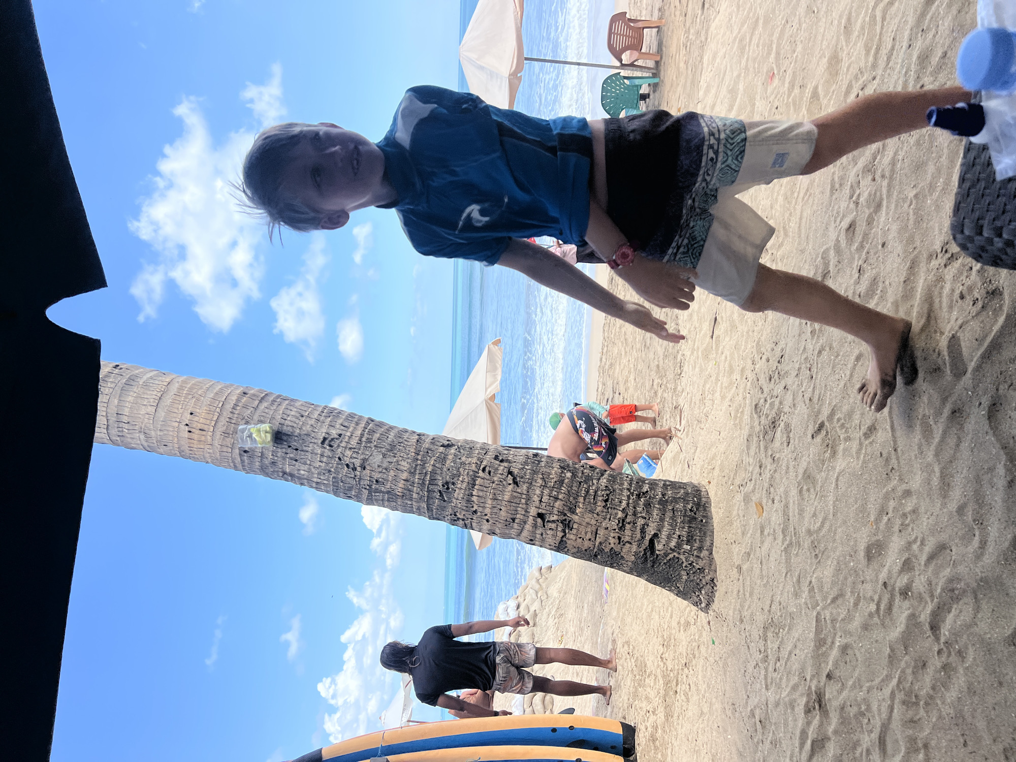 In Kuta, Indonesia, a young boy with blond hair is walking on a sandy beach. He is wearing a blue rash guard and patterned shorts. The boy is on the right side of the image, facing left and looking to the right. In the background, a man with long dark hair is standing near a surf board, facing away from the camera. Further back, two people are sitting on chairs under umbrellas near the ocean. A tall palm tree is prominently featured in the center of the image, leaning to the left. The sky is blue with scattered white clouds, and the ocean is visible in the background. The overall scene depicts a relaxed day at the beach.