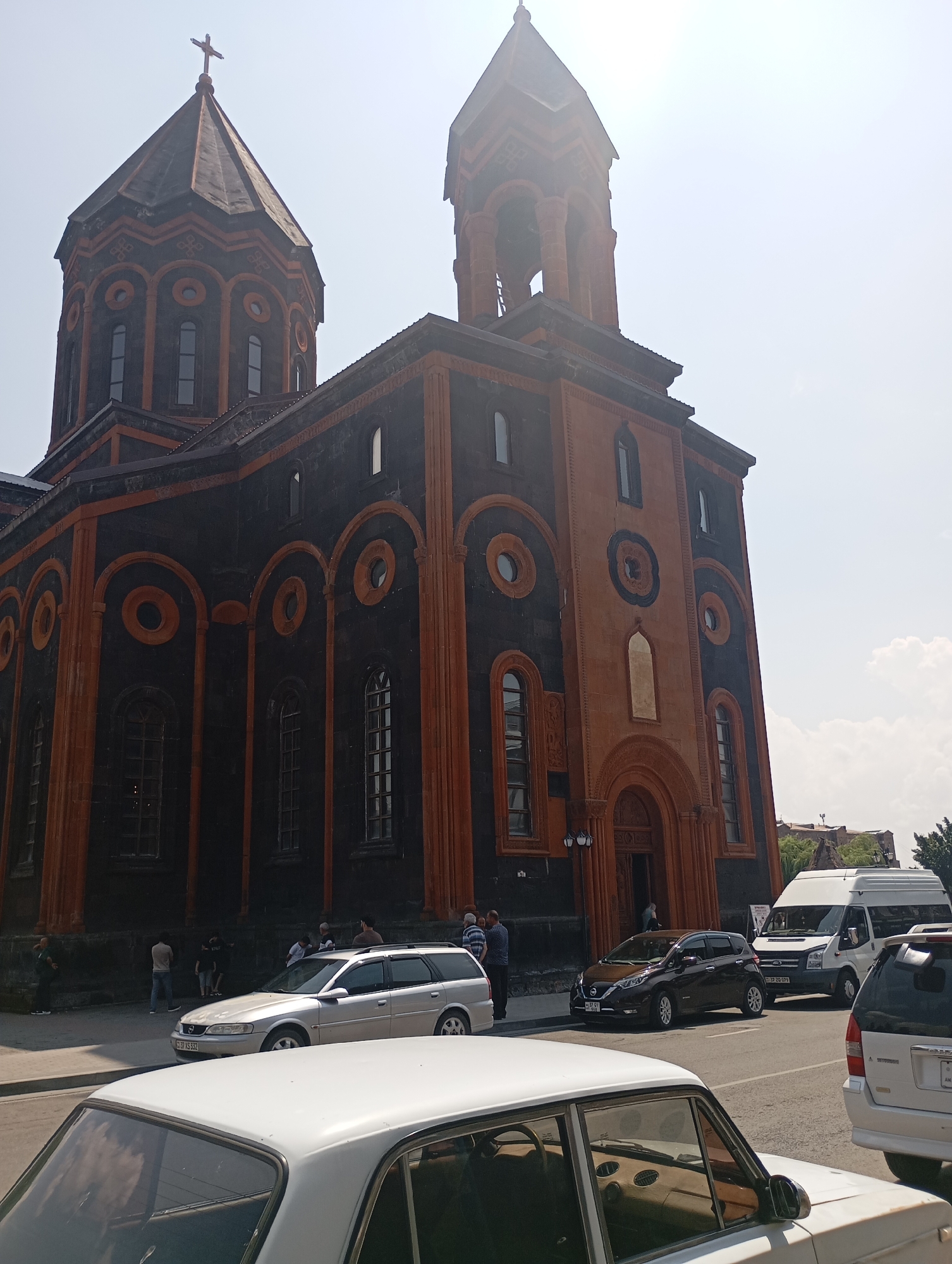 A wide shot captures the exterior of the Holy Mother of God Cathedral in Gyumri, Armenia, on a sunny day. The cathedral, a significant religious landmark, features a prominent bell tower and a large dome, both adorned with crosses. The building's facade is a striking combination of dark volcanic stone and reddish-orange brickwork, with arched windows and intricate decorative elements.
In the foreground, several cars are parked on the street. These include a white sedan, a silver station wagon, and a dark-colored hatchback. A white van is also visible. A few individuals are present near the cathedral entrance and on the sidewalk, some appearing to be tourists or worshippers. The street is paved, and there are some trees and buildings visible in the background. The sky is bright and clear, with a few white clouds. The overall scene depicts a typical day at a prominent Armenian church, with a mix of architectural grandeur and everyday urban life.