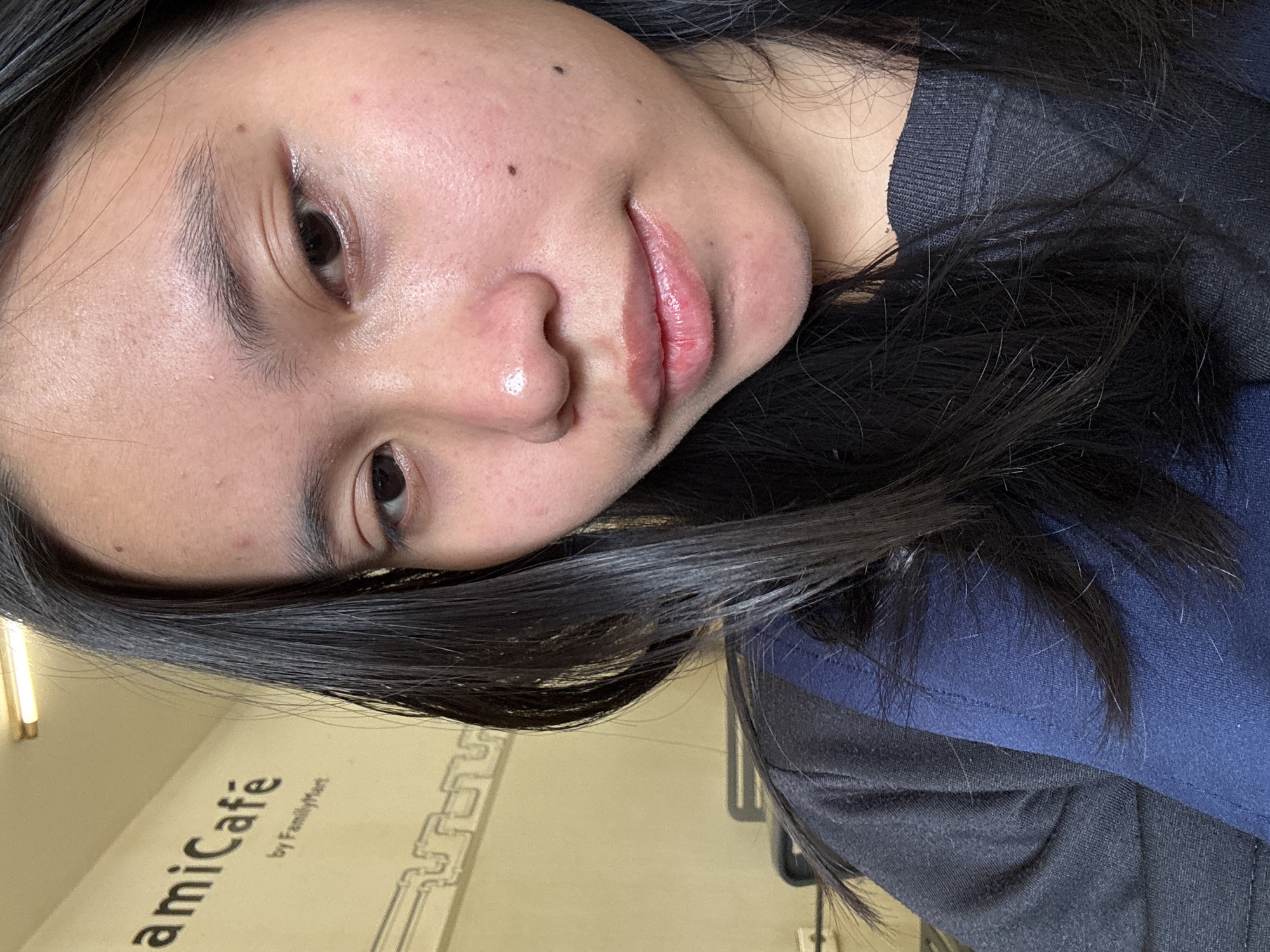 This is a close-up, indoor, selfie-style photograph of a young East Asian woman. The woman has long, dark, straight hair, and is looking slightly to her right. Her face is the primary focus, with her skin appearing to have some minor blemishes and redness. She is wearing a dark-colored sweatshirt with a blue panel across the shoulder and chest area. In the background, a wall with a sign that reads "amiCafe" and "by FamilyMart" is visible, suggesting the location is likely a FamilyMart convenience store cafe in East Jakarta, Indonesia. The lighting appears to be artificial, likely from overhead fluorescent lights, as indicated by the light source visible at the top of the frame.
