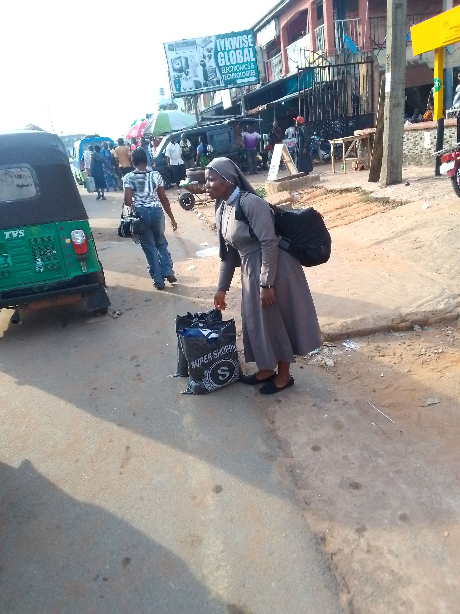 The image captures a street scene in Orlu, Nigeria. A Catholic nun wearing a gray habit and black shoes stands next to a black shopping bag labeled "Super Shopping" with a large "S" logo. She is bending down, seemingly adjusting items inside the bag. A black backpack is slung over her shoulder. To her left, a green tricycle taxi labeled "TVS" is visible. In the background, a crowd of people walks along the street. Overhead is a billboard advertising "IYKWISE GLOBAL ELECTRONICS & TECHNOLOGIES." The street is paved and appears worn, with some dirt and debris. Further back, a building is visible with a metal gate and an elevated platform where people are sitting. A yellow sign with the word “olo” and a concrete pole are next to the building.