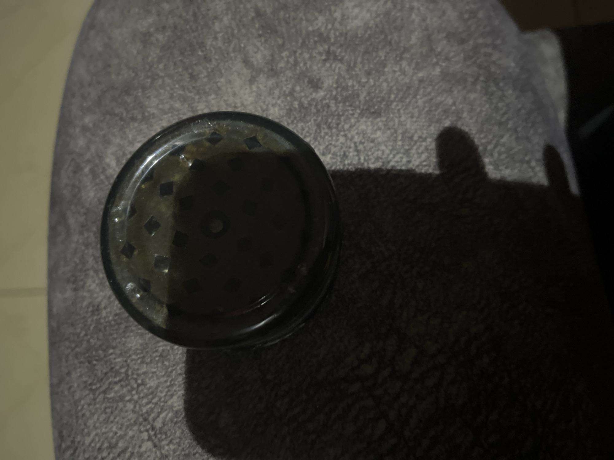 A dark, circular object, possibly a container or grinder, sits on a textured, grey surface, which appears to be fabric or upholstery. The object is dark and slightly transparent, with a patterned interior visible through its surface. Light casts a shadow from the object onto the surface, and a hand or finger is partially visible in the lower right corner, creating a distinct shadow. The background is blurred and appears to be a light-colored tiled surface, suggesting an indoor setting. The scene suggests a close-up, still life composition. The location in Jos, Nigeria, is not visually indicated by any elements within the image.
