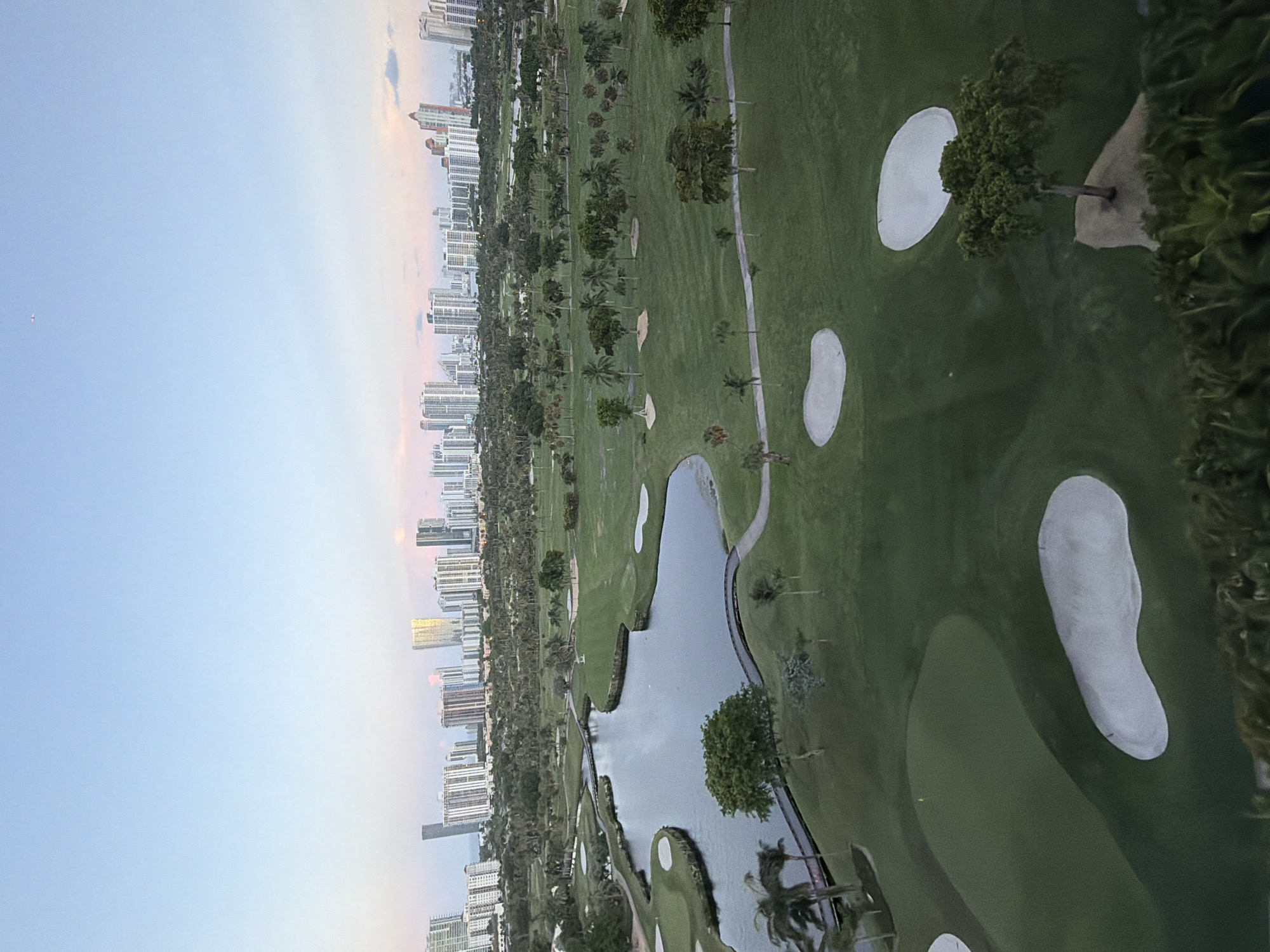 The image shows an aerial view of a golf course in Aventura, Florida, United States of America. The golf course features several holes, sand traps, a body of water, and bridges. In the background, a cityscape with numerous high-rise buildings is visible, stretching across the horizon. The sky is clear with a light blue hue, suggesting it might be early morning or late evening. There are no people visible in the scene. The overall impression is one of a well-maintained golf resort adjacent to a densely populated urban area.